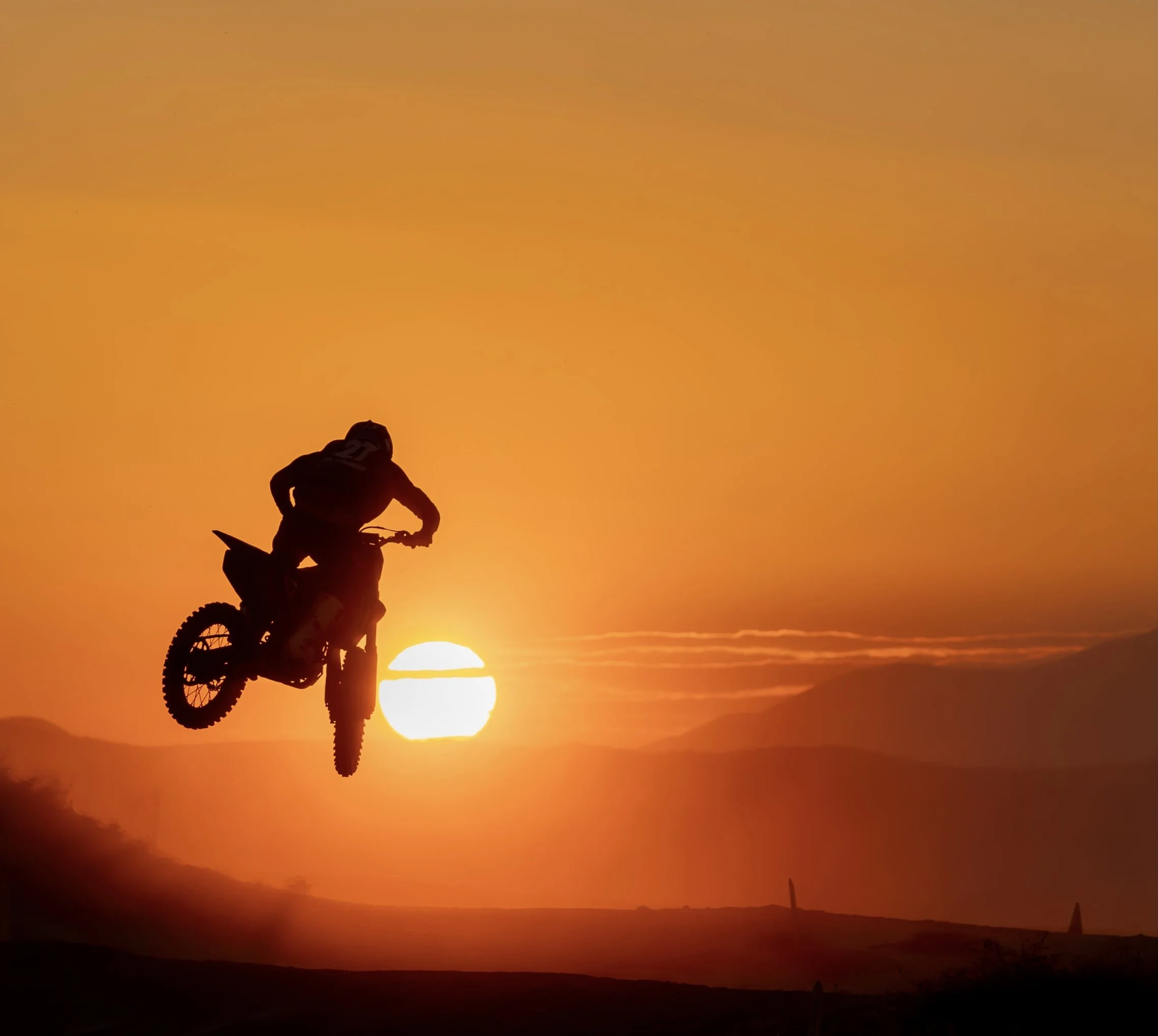 Persona haciendo salto con motocicleta durante atardecer, con el sol en el horizonte y montañas en el fondo.