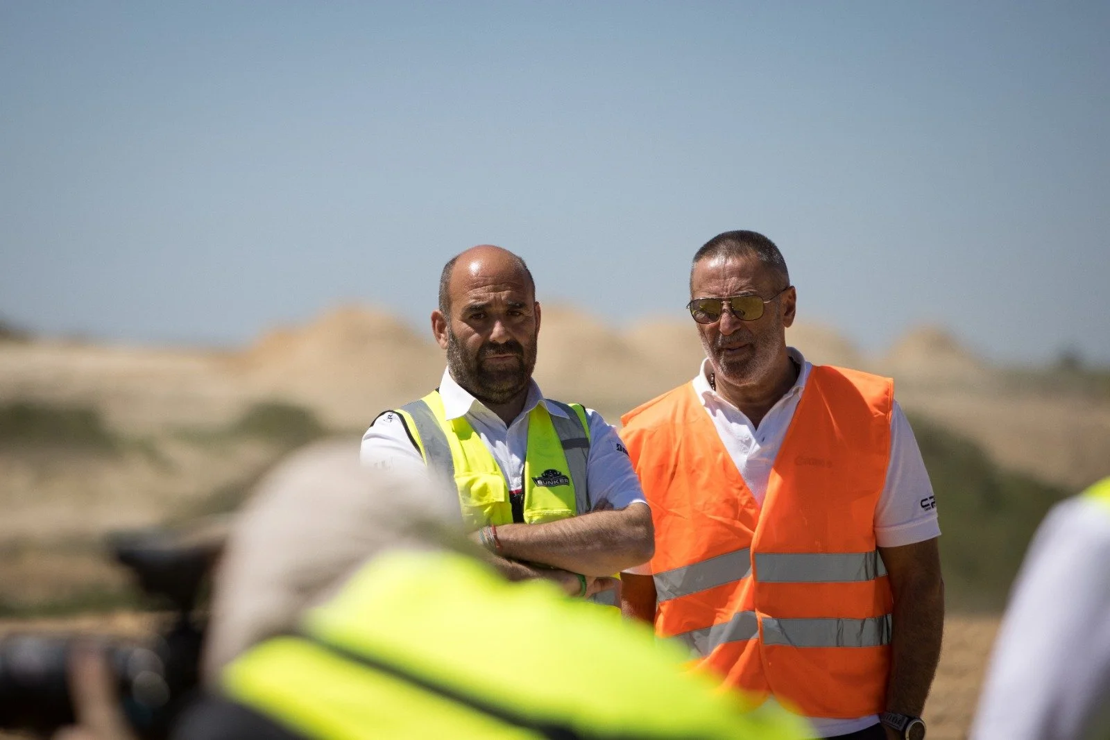 Dos hombres con chalecos de seguridad en un entorno exterior con colinas en el fondo.