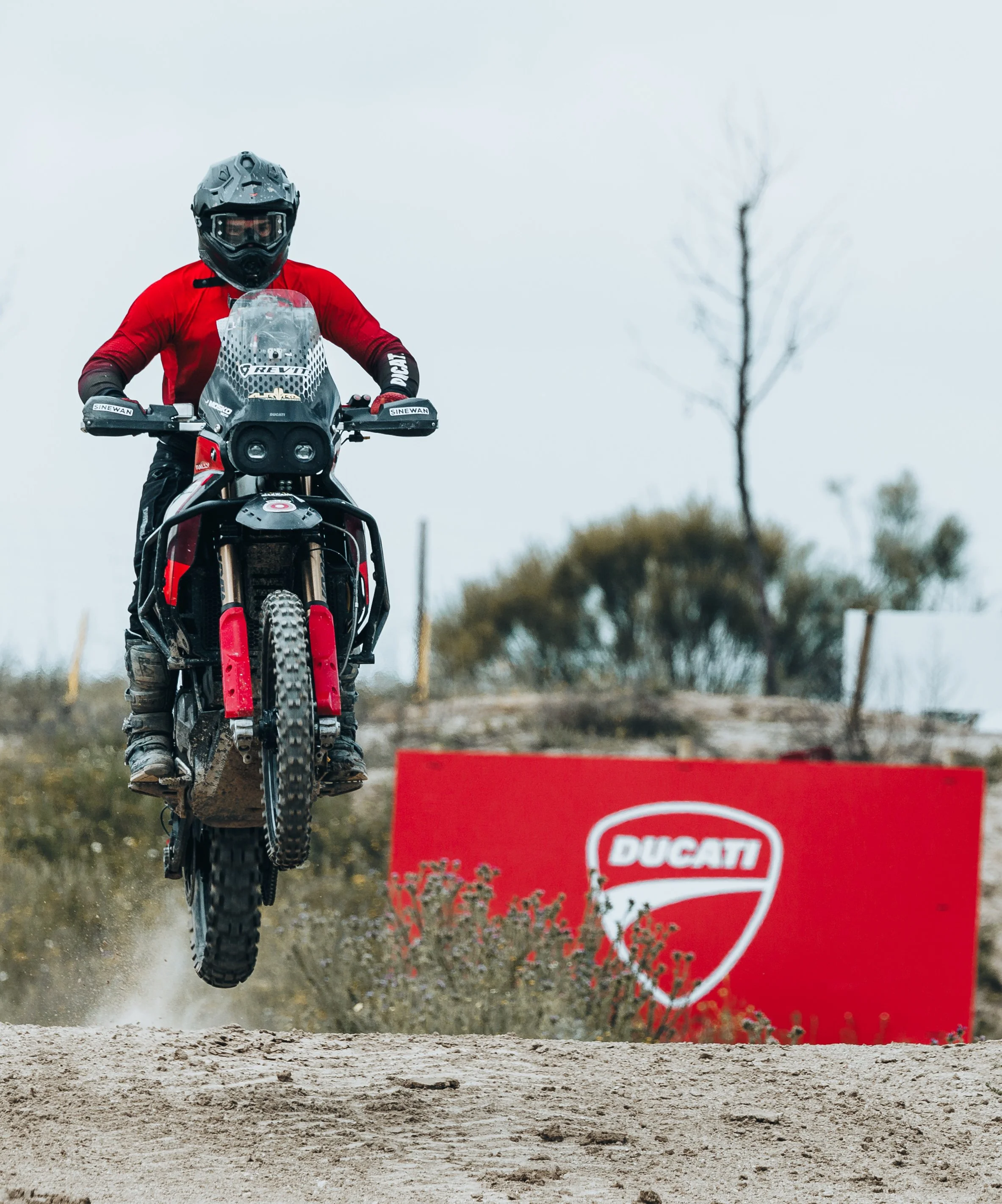 Persona montando una motocicleta de deporte de color negro y rojo en un terreno de tierra, con un letrero grande de Ducati al fondo.