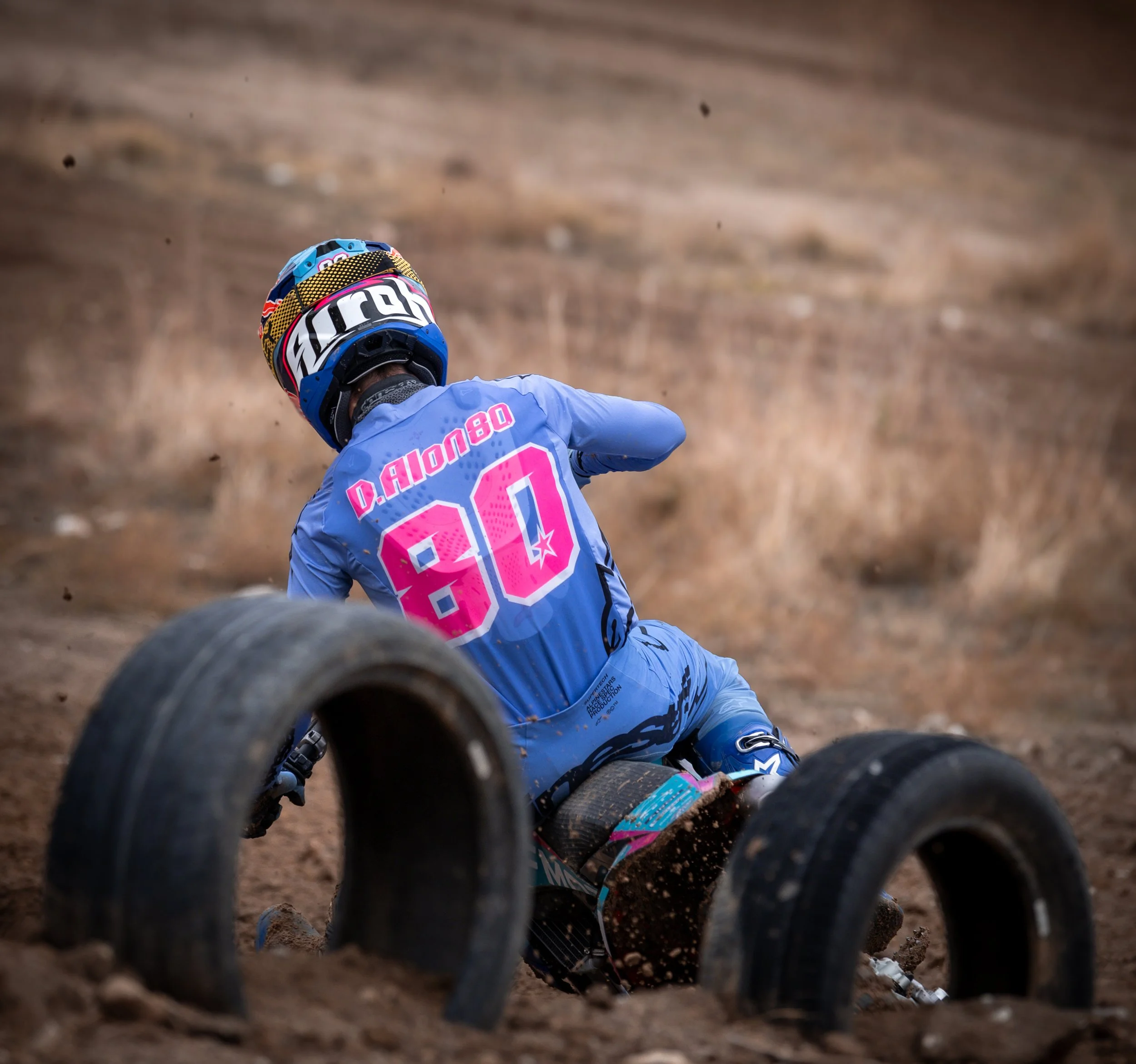 Un motociclista en uniforme azul con el número 80 y el nombre D. Alonzo en la espalda, cayendo en un camino de tierra con neumáticos. Lleva casco con varios colores y está en medio de una caída o accidente durante una carrera de motocross.