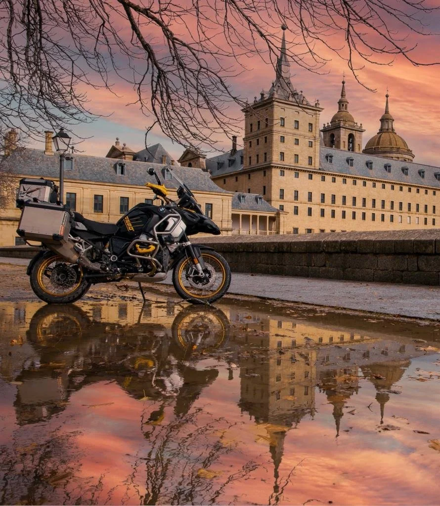 Motocicleta negra en charco reflejando un castillo con cúpulas doradas bajo un cielo rosa y nublado, con ramas de árbol en primer plano.