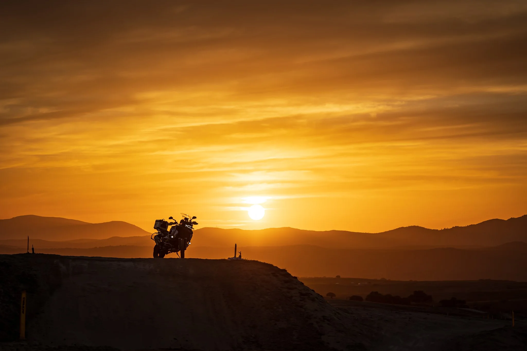 Motocicleta en un paisaje al atardecer con montañas en el horizonte y un cielo con tonalidades doradas.