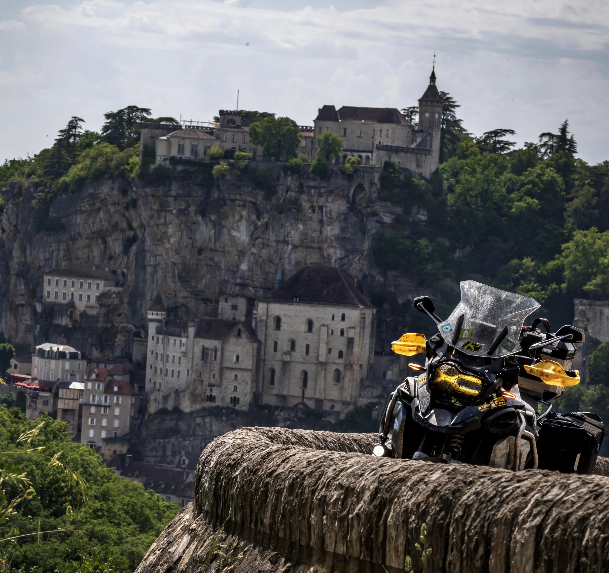 Vista de un castillo en una colina con un arco y árboles verdes, una motocicleta negra y amarilla en primer plano sobre un muro de piedra.