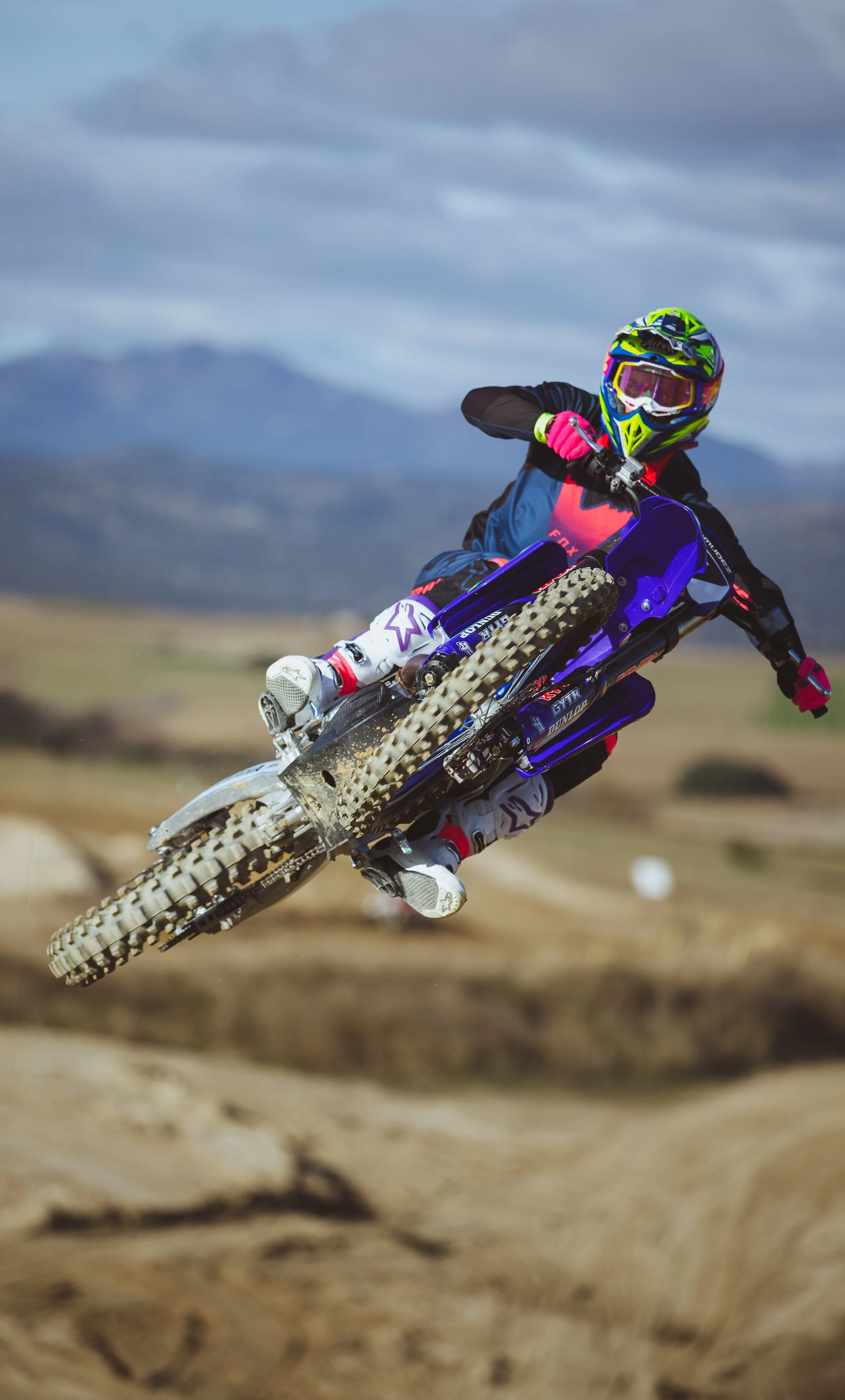 Un motociclista realizando un salto en una pista de tierra para motocross, llevando equipo de protección y casco de colores brillantes.