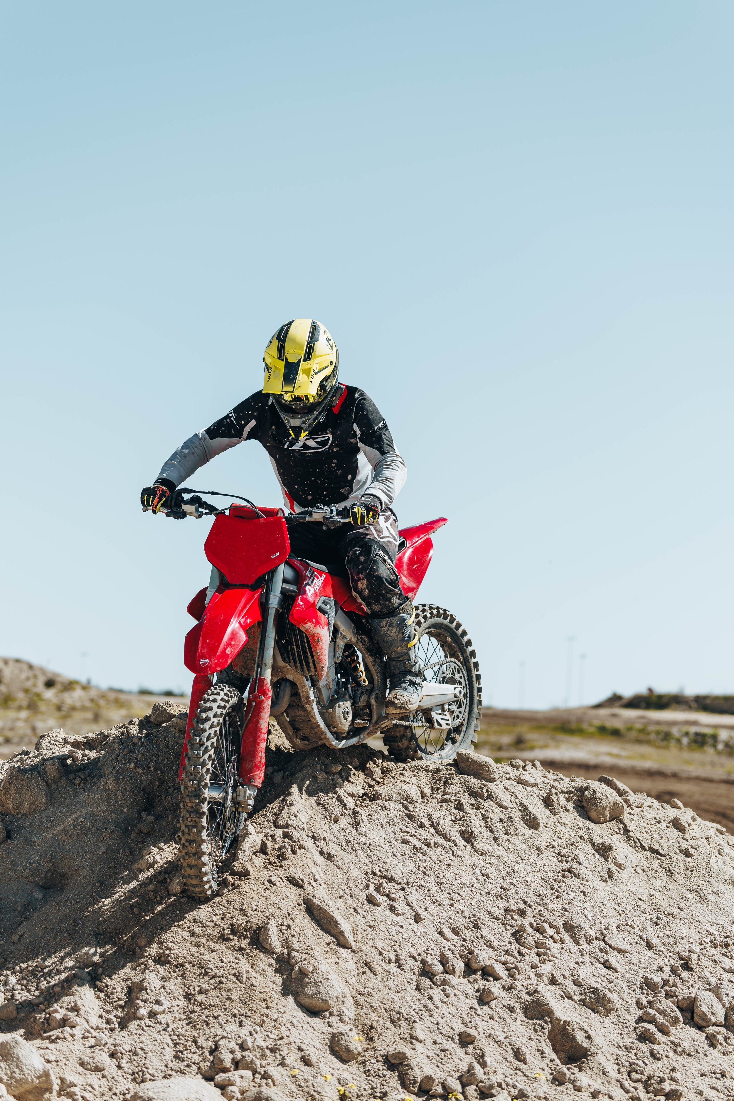 Persona montando una moto de cross roja en un terreno de arena y rocas bajo un cielo despejado.