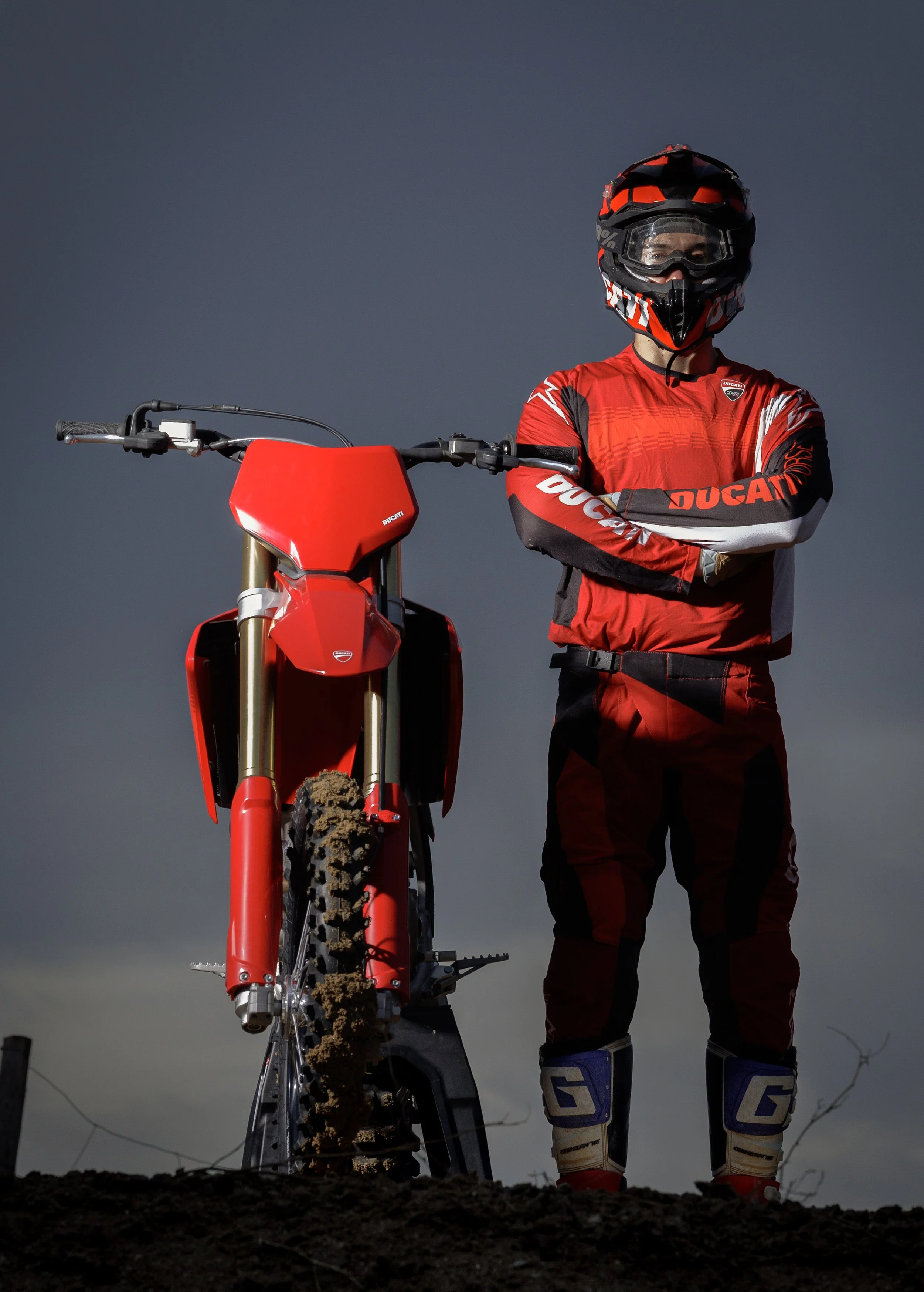 Persona con equipo de motocross, casco y botas, de pie junto a una motocicleta de dirt bike roja, en un paisaje con cielo oscuro o nublado.