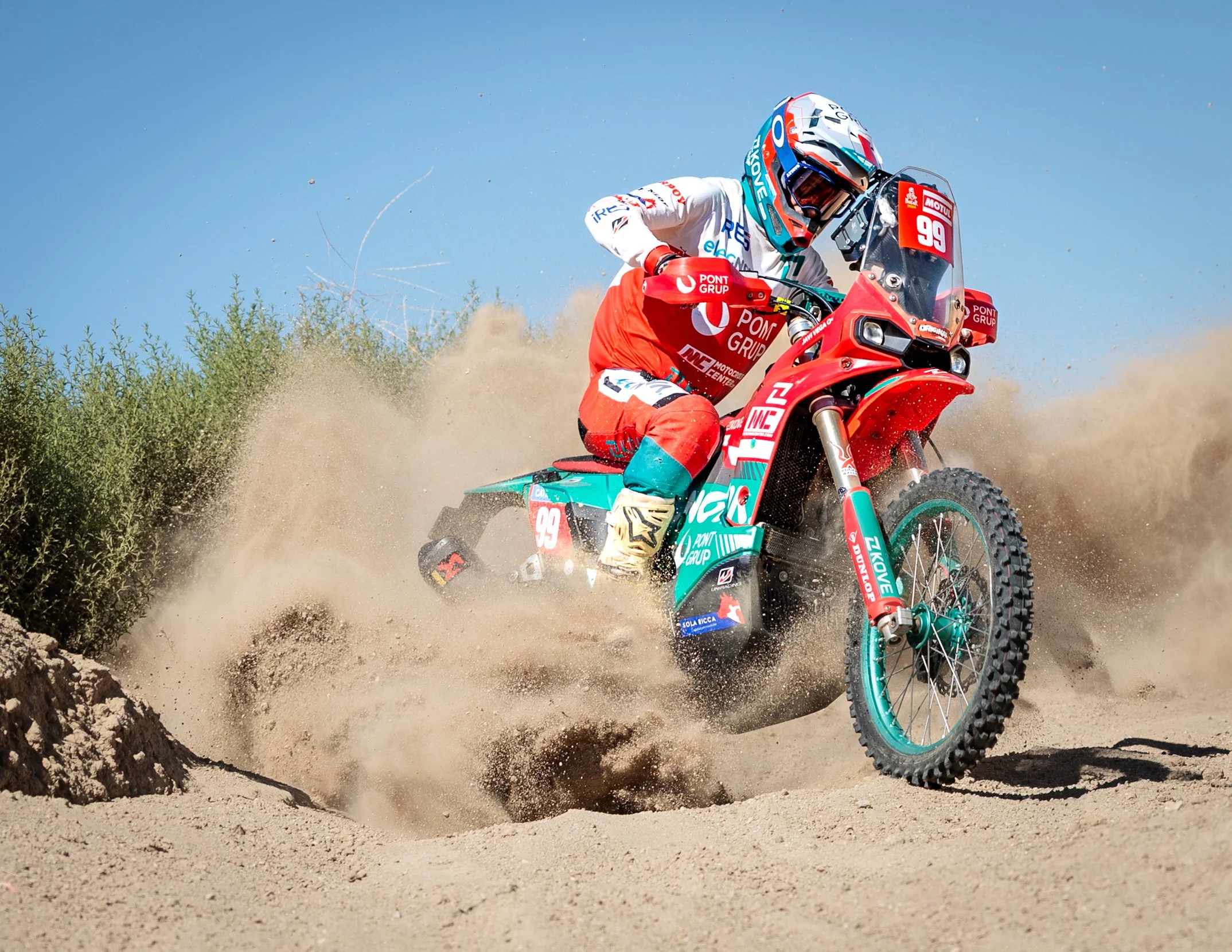 Motociclista en acción en un terreno de arena, con polvo levantado y vegetación en el fondo, vestido con equipo de protección y uniforme de competición