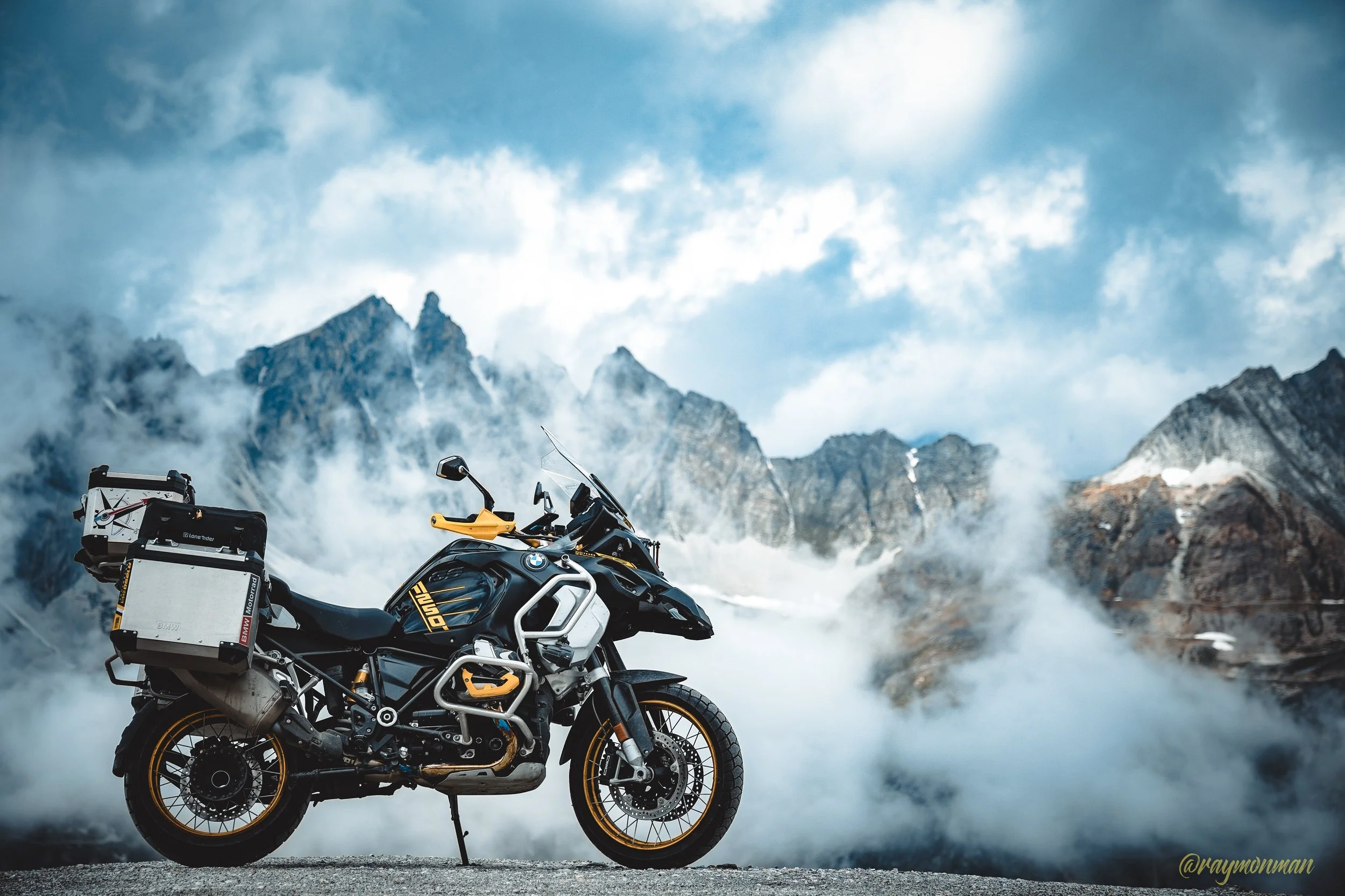 Motocicleta BMW Adventure en una carretera montañosa con picos nevados y nubes.