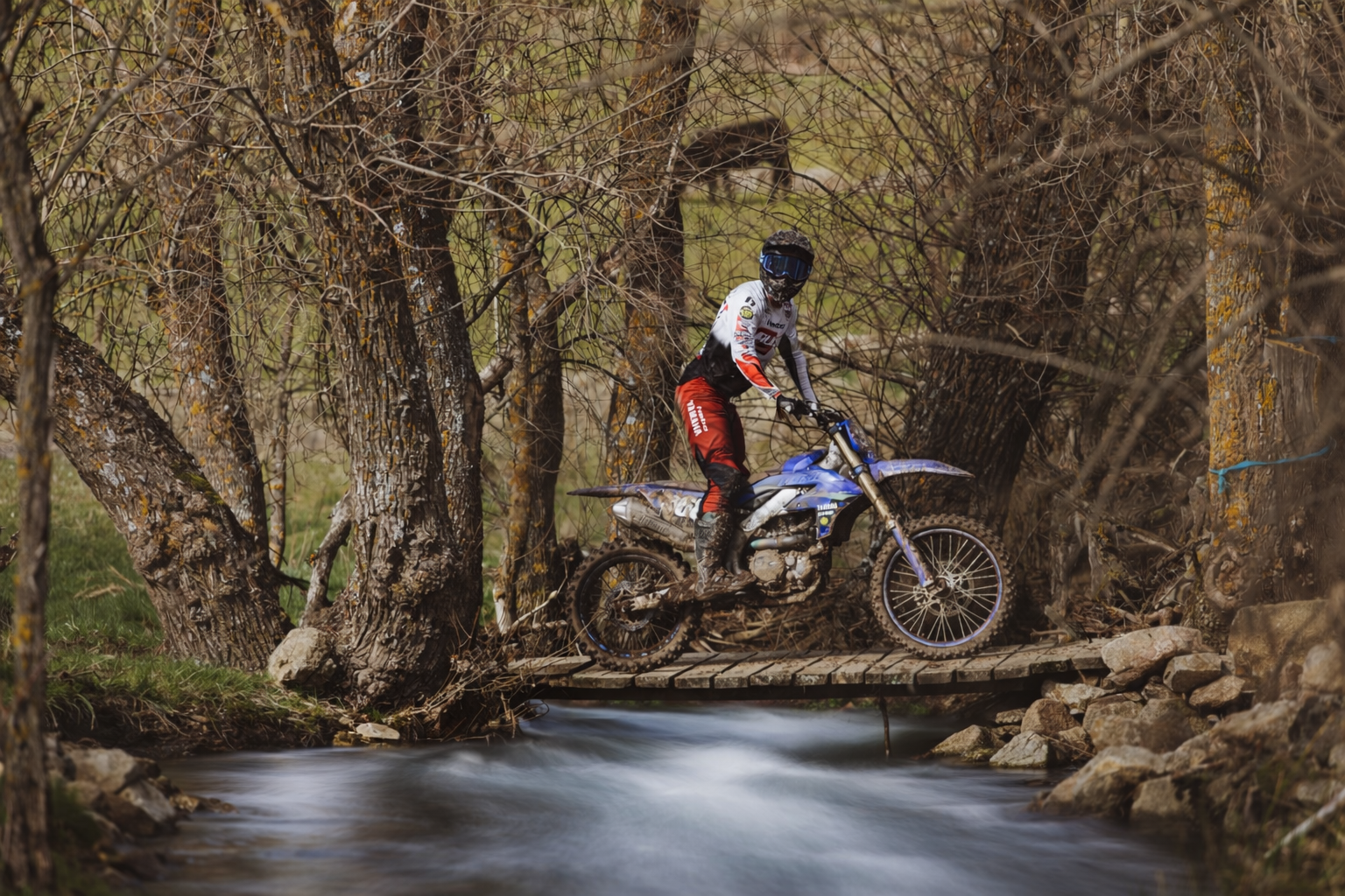 Persona montando una motocicleta de enduro en un puente de madera sobre un río en un entorno natural con árboles y rocas.