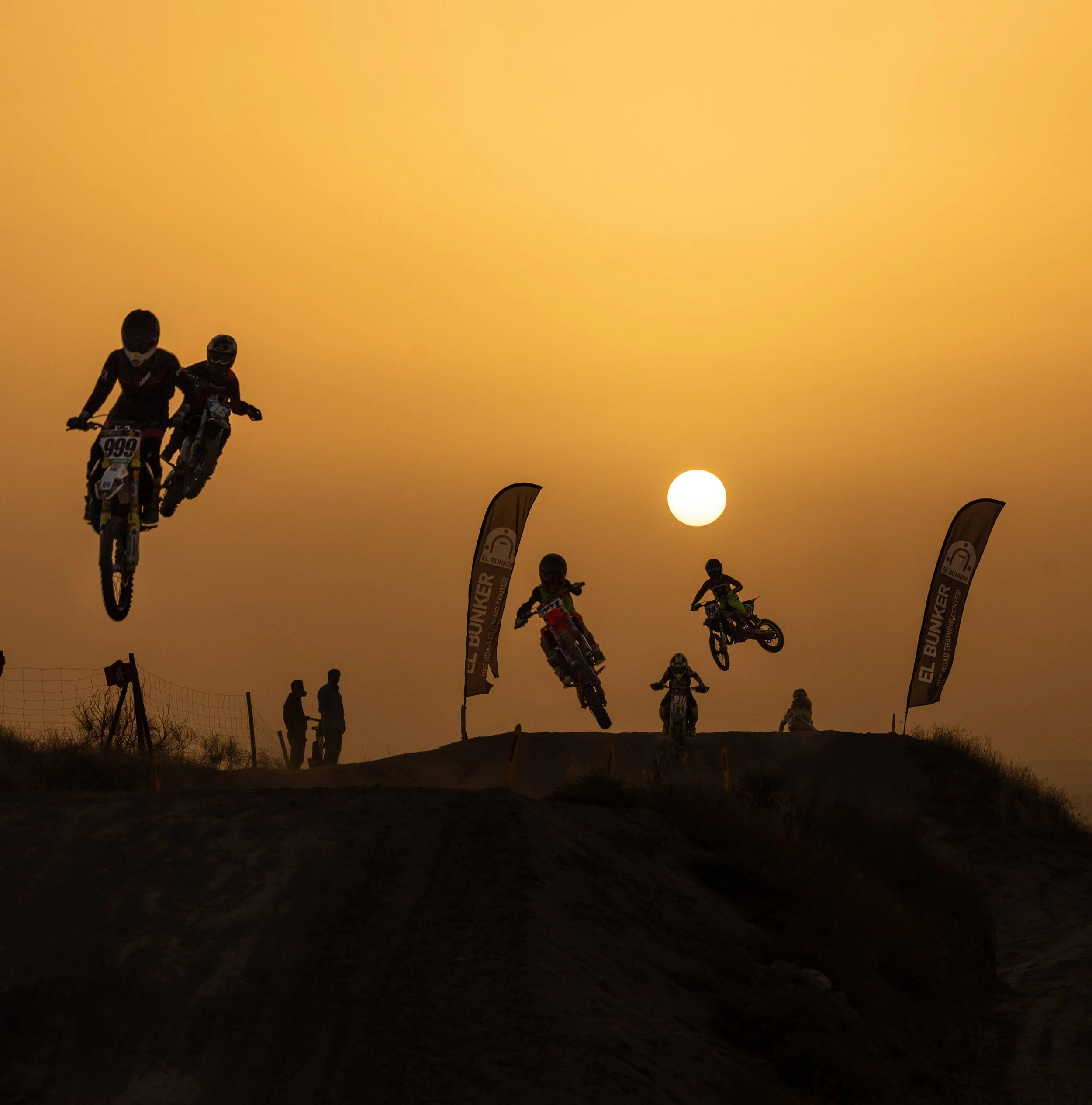 Fotografía de ciclistas en salto durante una competición al atardecer, con un sol visible en el cielo y banderas que dicen 'El Bunker'.