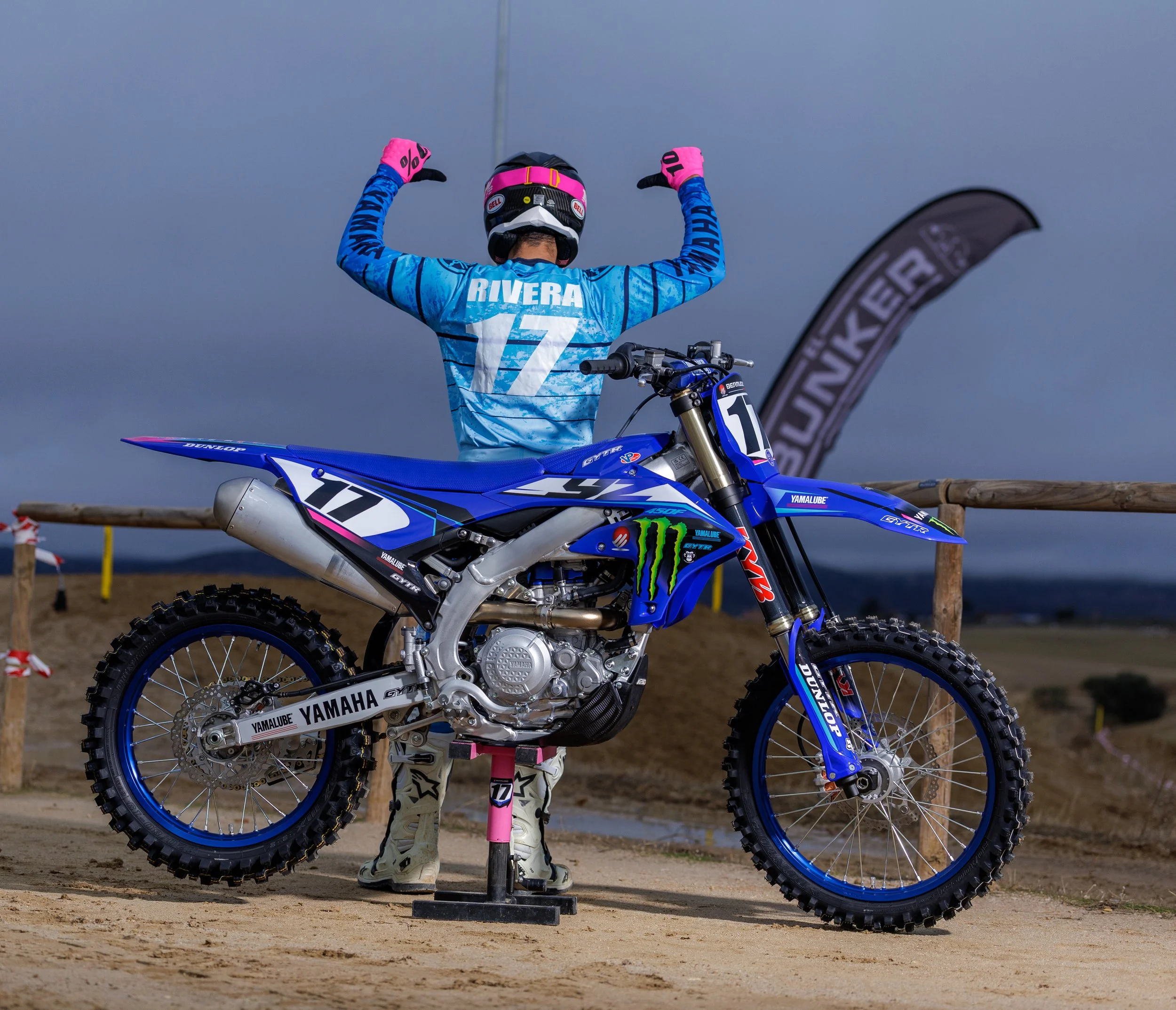 Un piloto de motocross de espaldas, vistiendo un jersey azul con el nombre Rivera y el número 17, levantando los brazos en señal de victoria frente a una moto azul de carreras, en un terreno fuera de pista con cielo nublado.