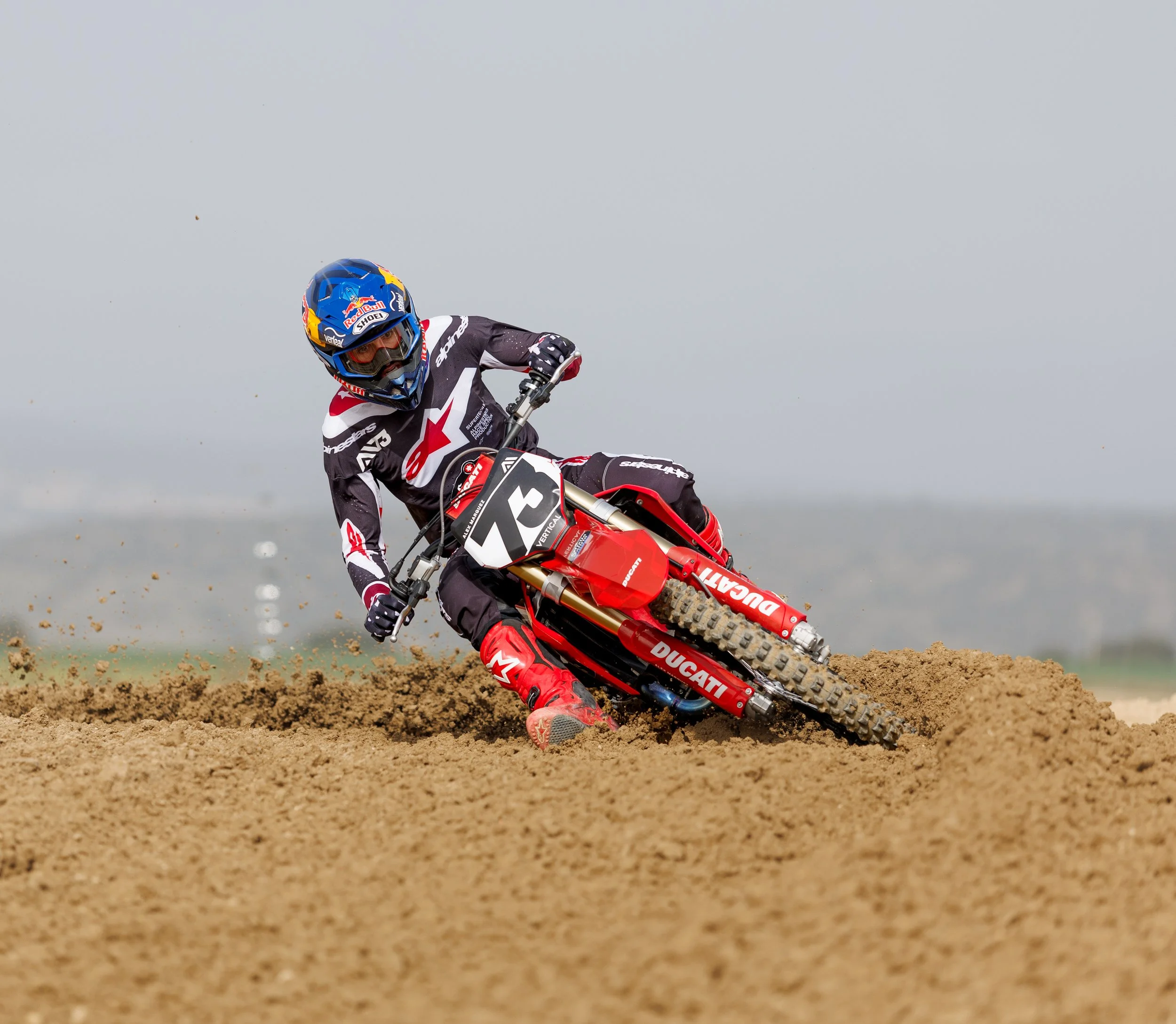 Motociclista en una carrera de motocross en tierra, inclinado en una curva, con casco azul y ropa de protección negra, roja y blanca.