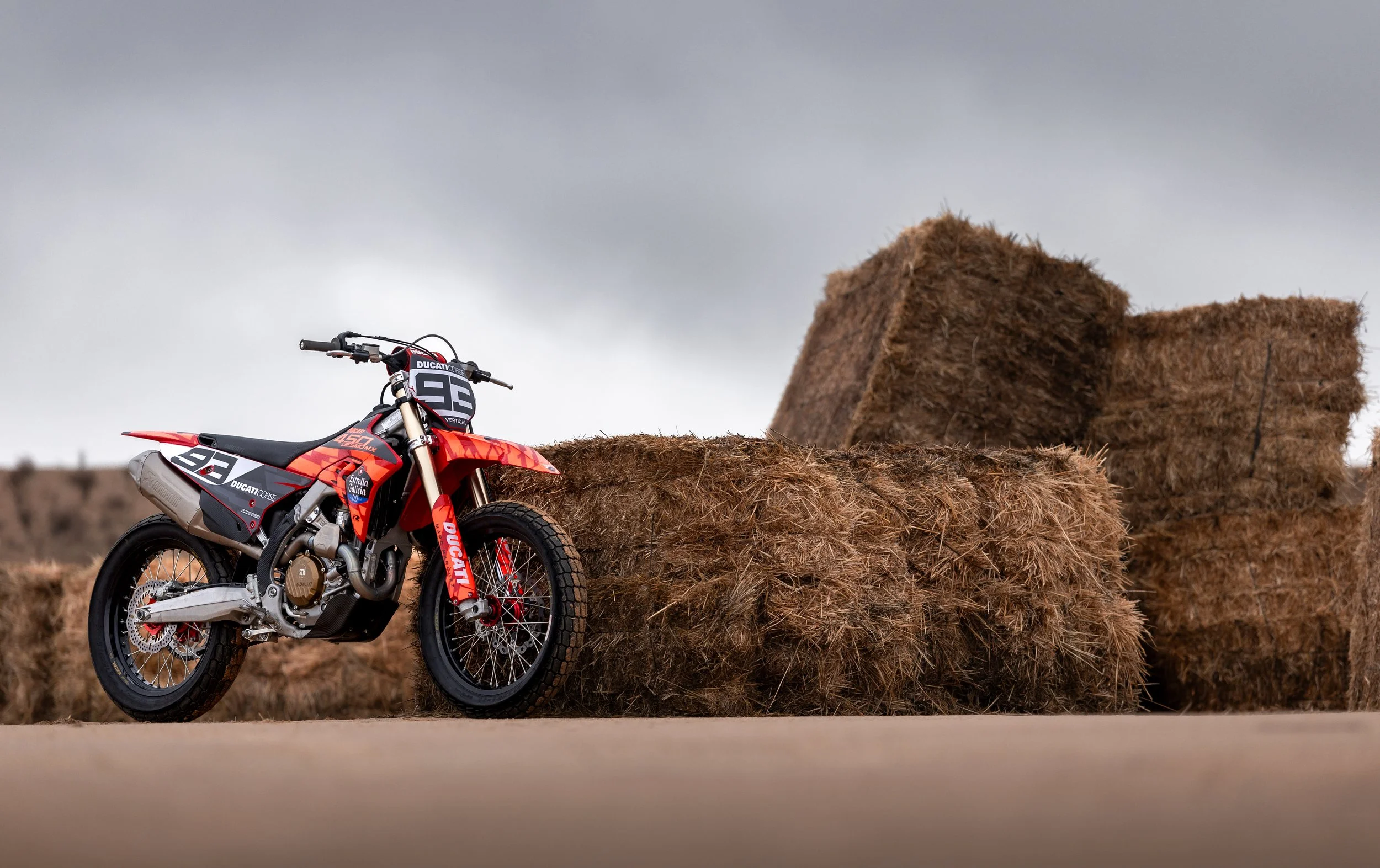 Motocicleta de carreras roja junto a pacas de heno en un campo con cielo nublado.