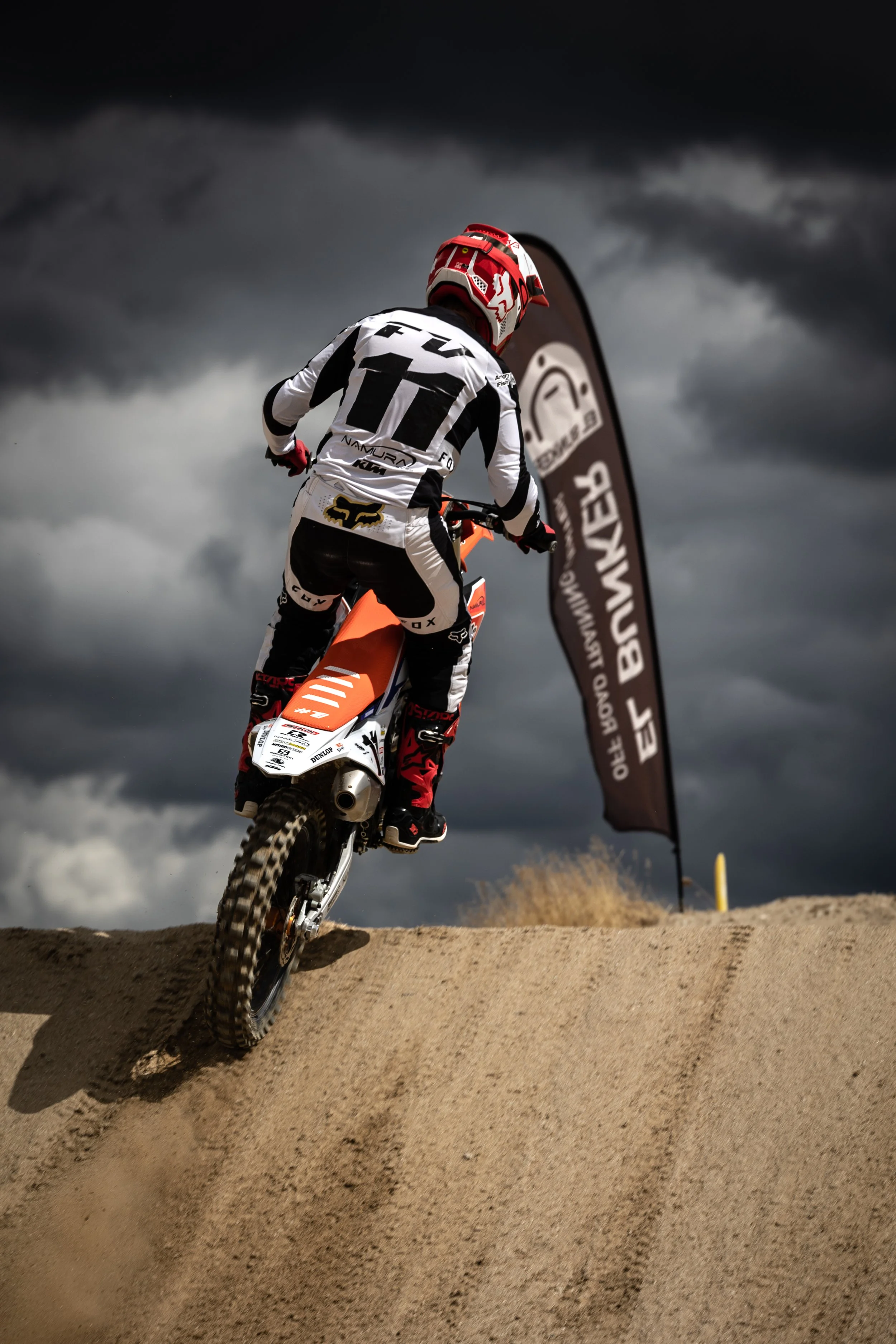Motociclista en un terreno arenoso bajo cielo nublado, con bandera de evento y vestimenta de protección.