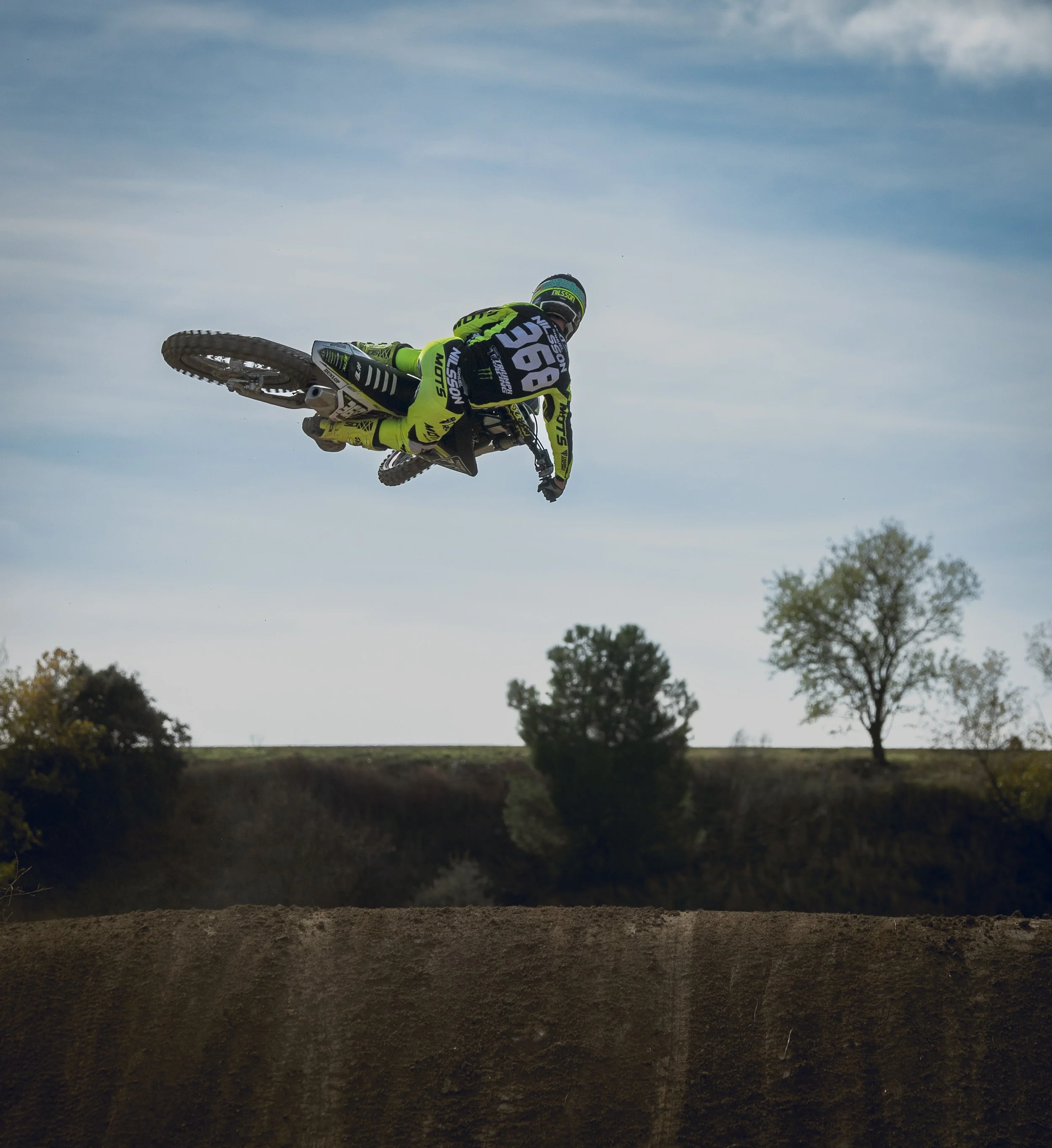 Jinete de motocross en el aire sobre un salto en el campo, con cielo y árboles de fondo.