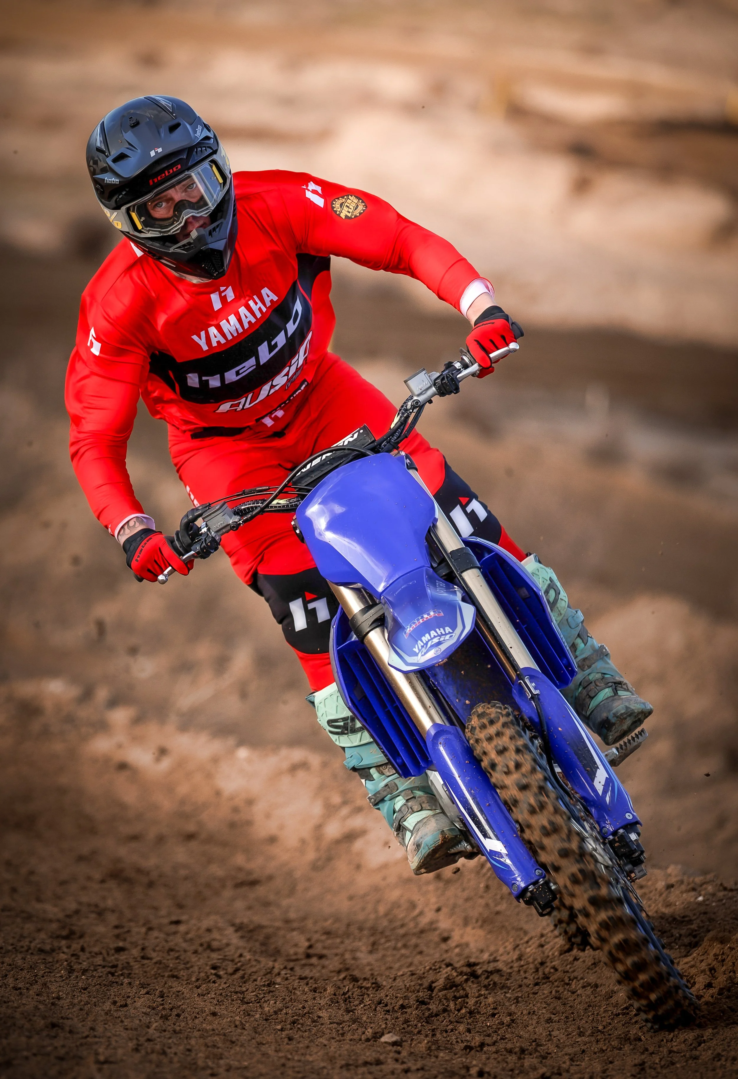 Un motociclista vestido con un traje rojo y un casco negro, montando una motocicleta azul en un terreno de tierra.