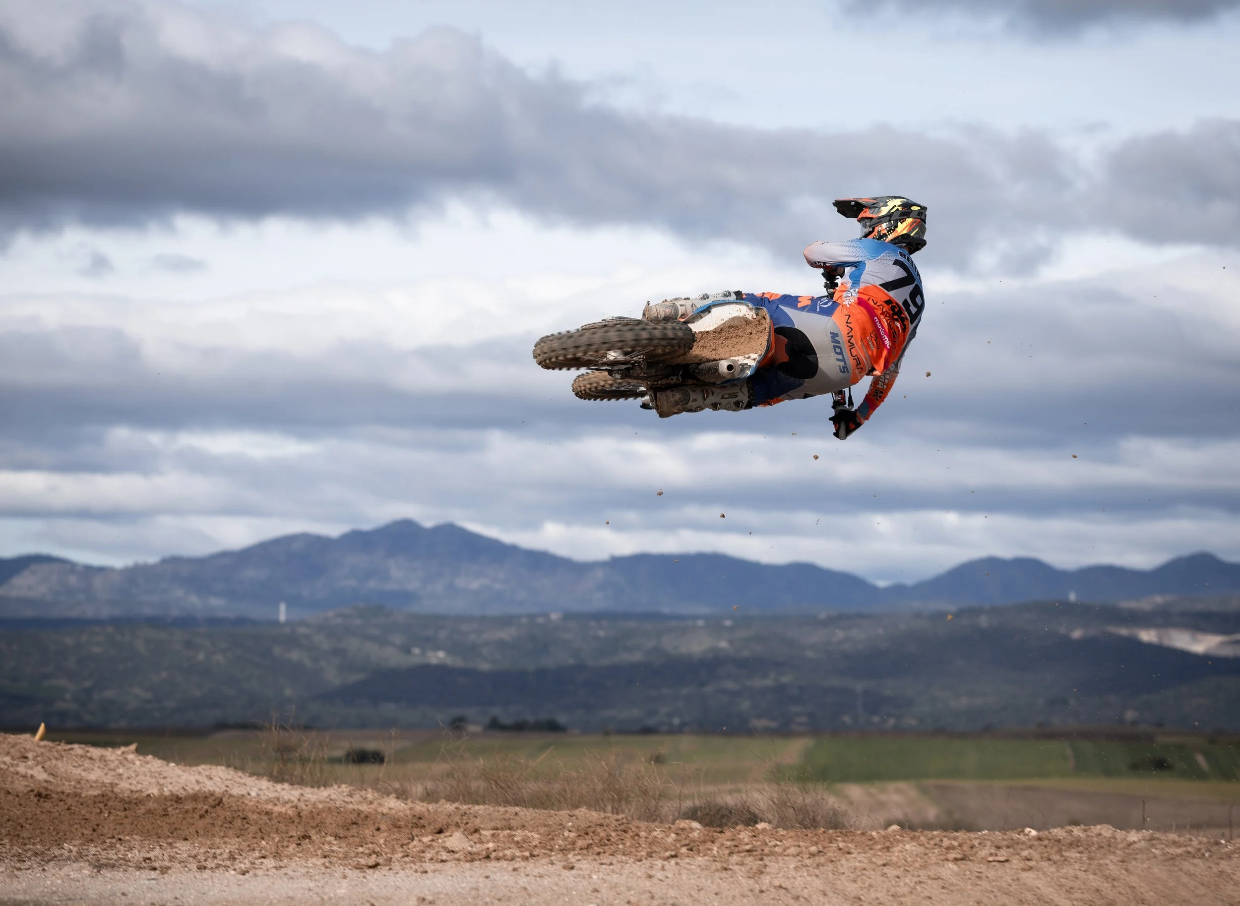 Un motociclista de motocross en medio de un salto en un circuito al aire libre con montañas y cielo nublado en el fondo.