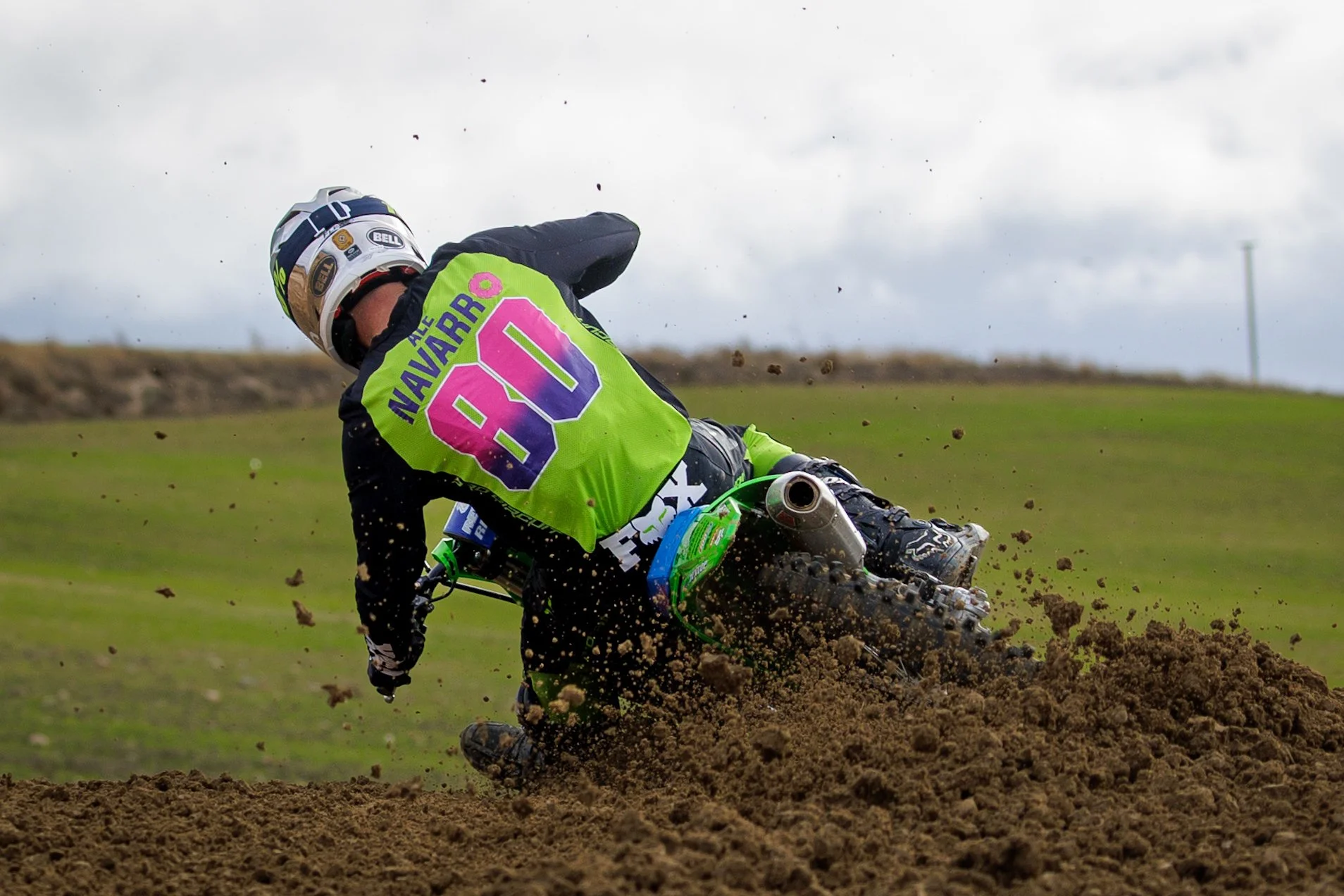 Un piloto de motocross en una curva de tierra, lanzando tierra y polvo con su motocicleta, vistiendo un traje negro y un casco gris con detalles oscuros. Lleva un chaleco verde con letras moradas y rosas que dice 'AL NAVARRO 80'