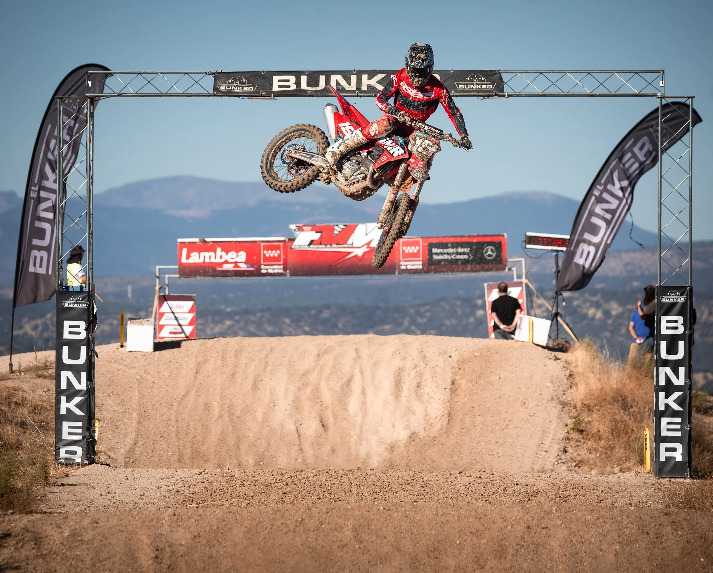 Un motociclista en salto en una carrera de motocross en un circuito al aire libre con montañas al fondo.