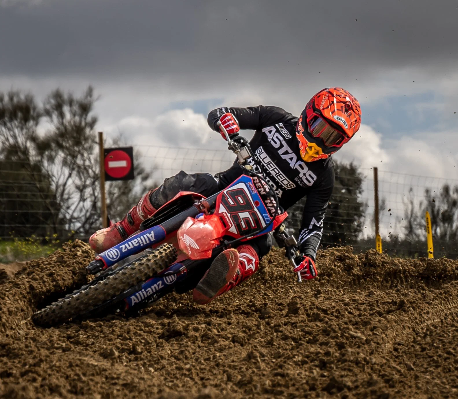 Motociclista en una pista de tierra, inclinándose en una curva, con fondo de árboles y cielo nublado