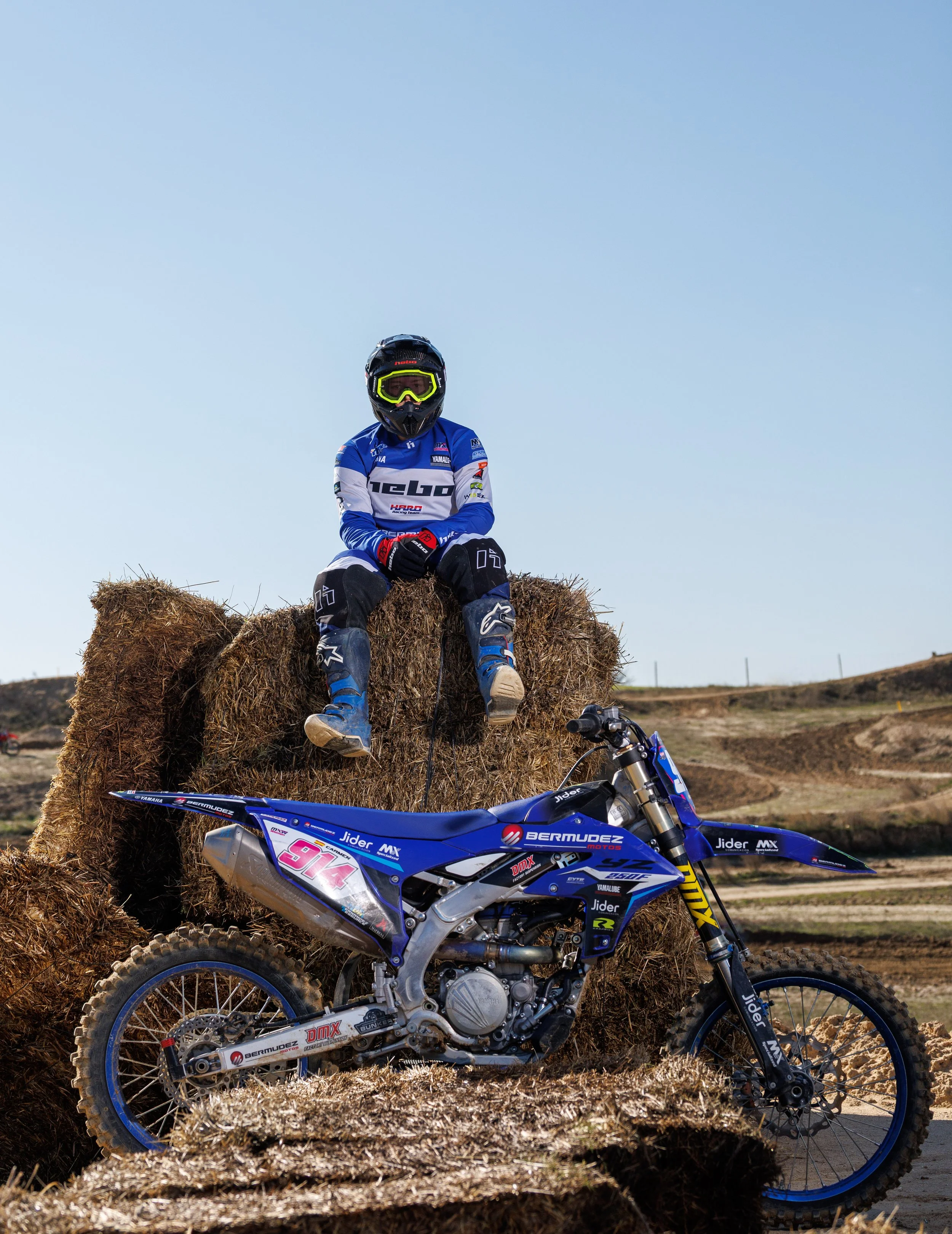 Un niño con equipo de motocross sentado sobre un montón de paja junto a una moto de dirt bike en un campo de tierra.