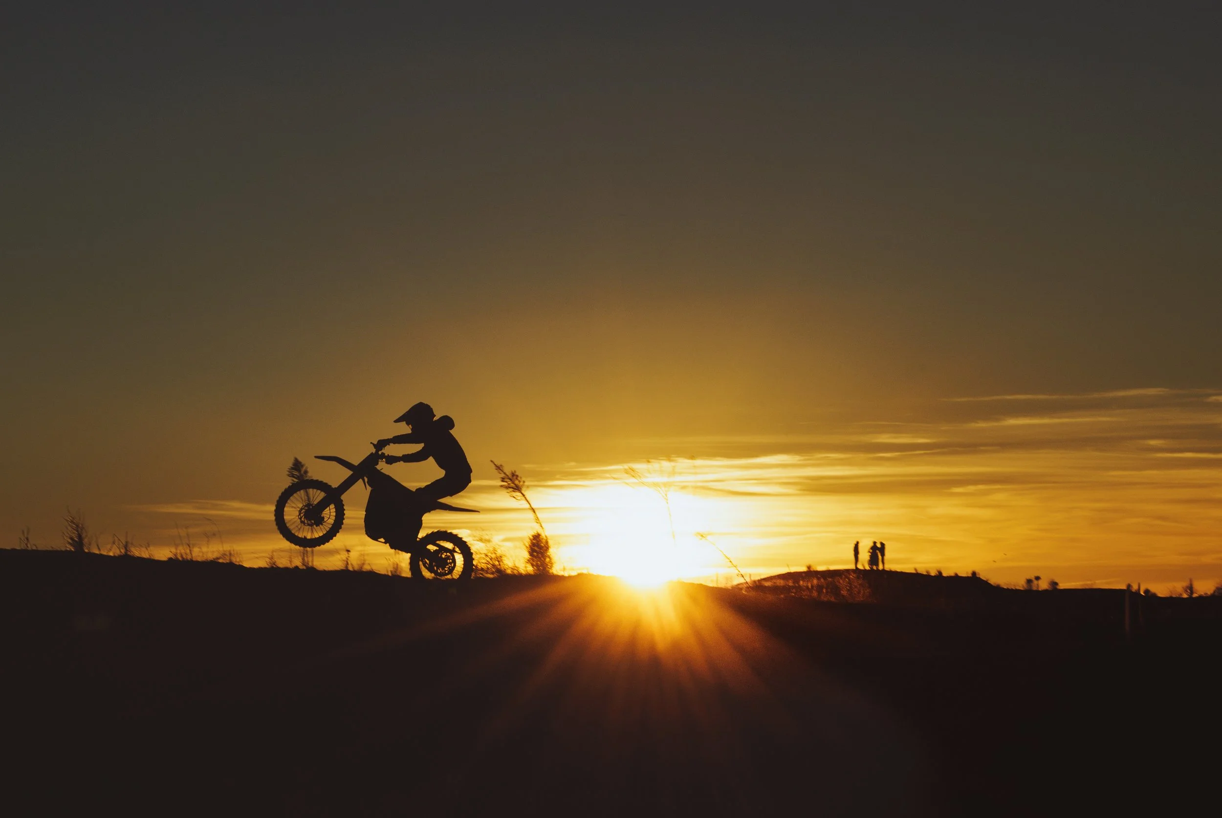 Persona realizando trucos en bicicleta durante un atardecer, con el sol en el horizonte y dos personas en la distancia.