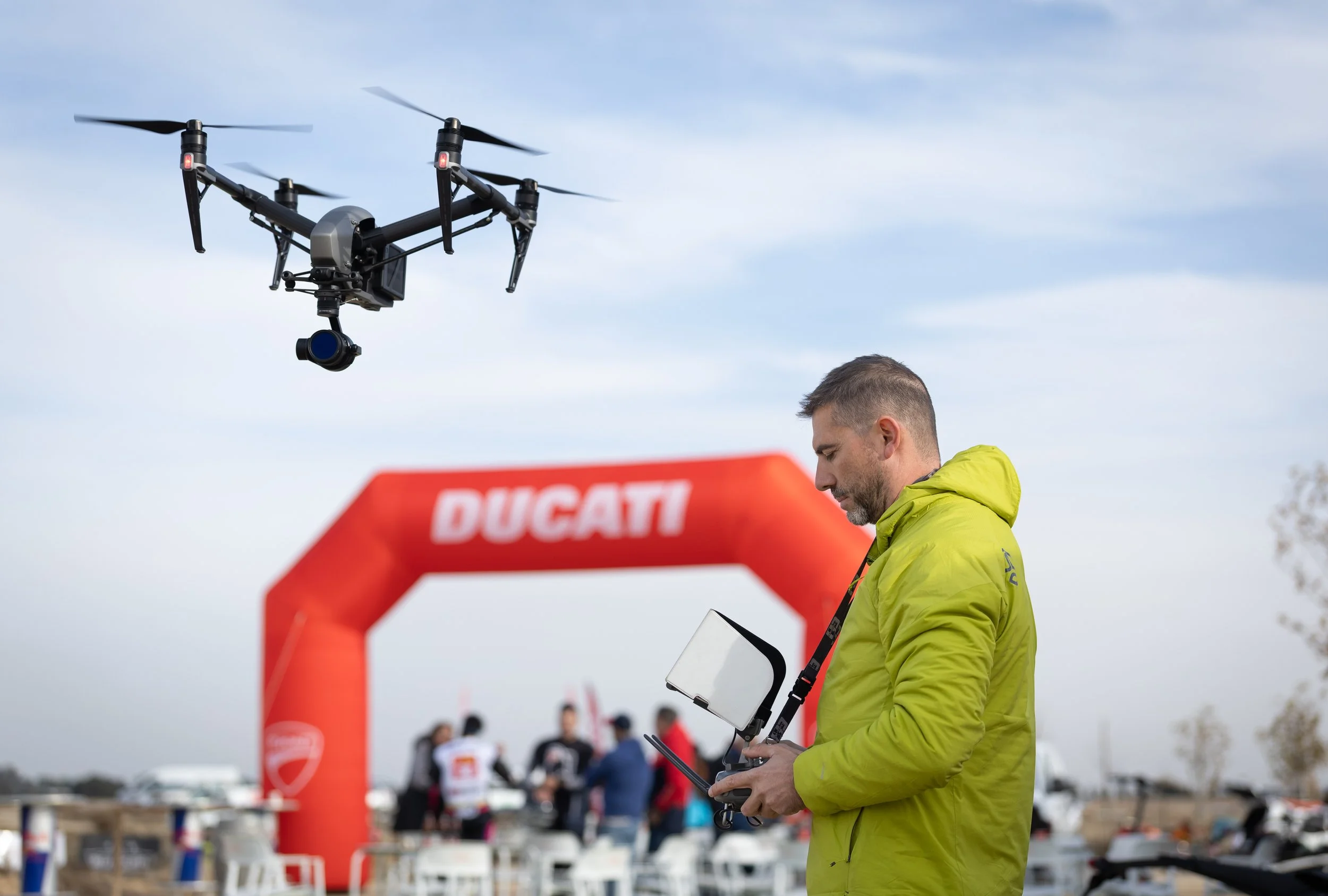Hombre con chaqueta verde controlando un dron en un evento de carreras de drones, con arco inflable de Ducati en el fondo y varias personas en el área.