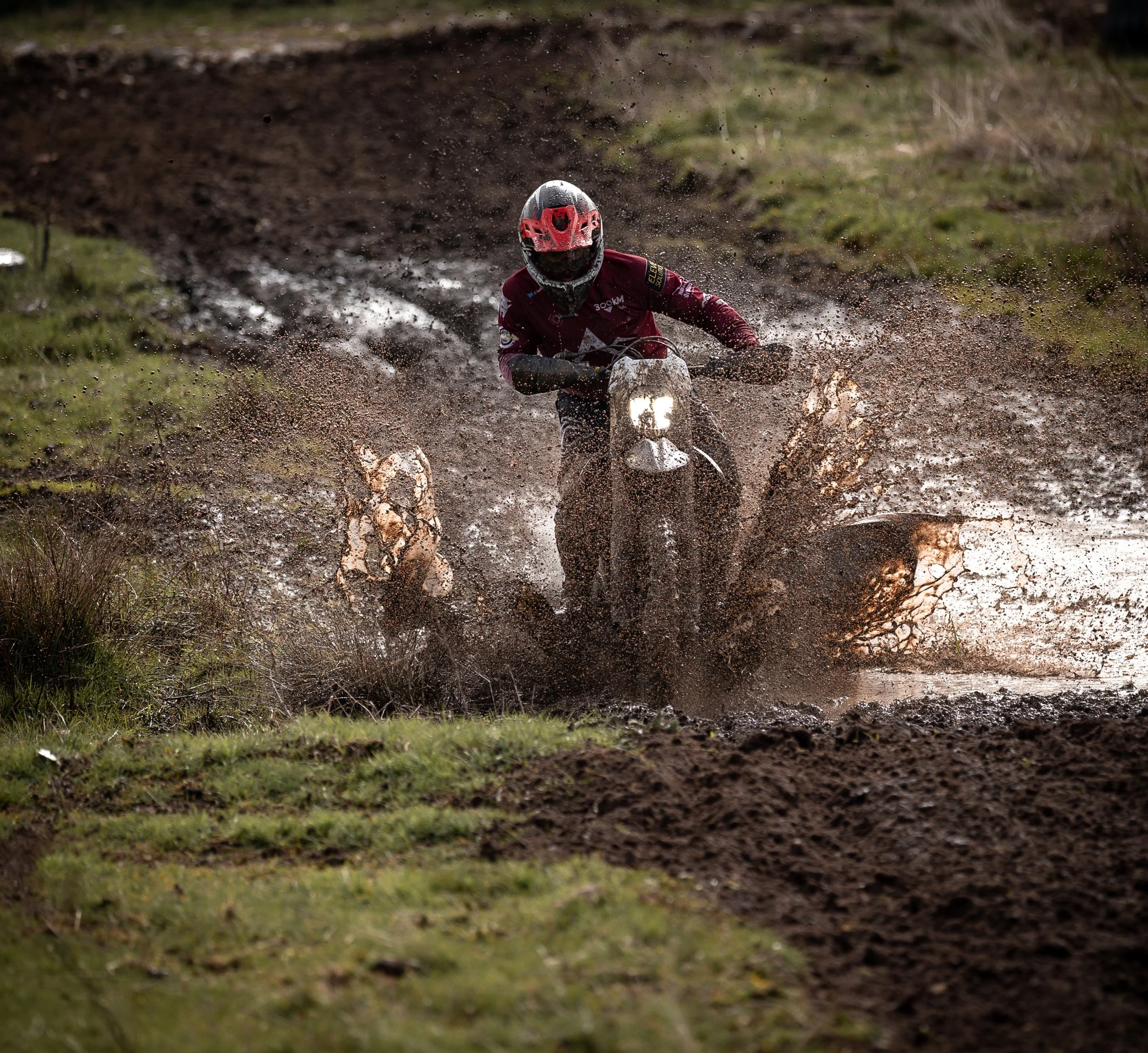 Una persona montando una motocicleta de enduro en un terreno fangoso, con agua y barro salpicándose mientras avanza.