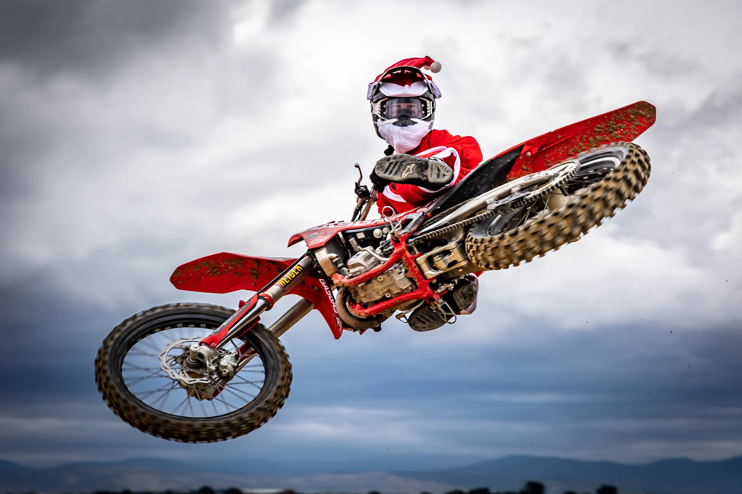 Un motociclista vestido con equipo de protección, incluyendo casco, gafas y ropa de color rojo, haciendo una acrobacia aérea en una moto de dirt bike de color rojo, con un cielo nublado de fondo.