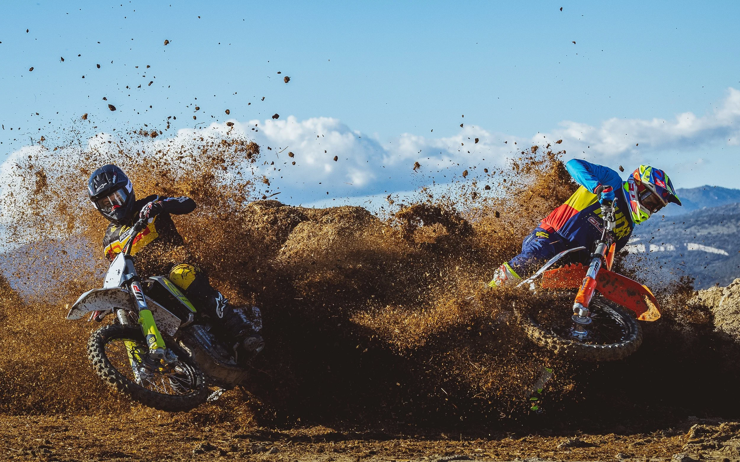 Dos motociclistas compitiendo en una carrera de motocicletas en terreno arenoso, con explosiones de polvo y tierra en el aire.