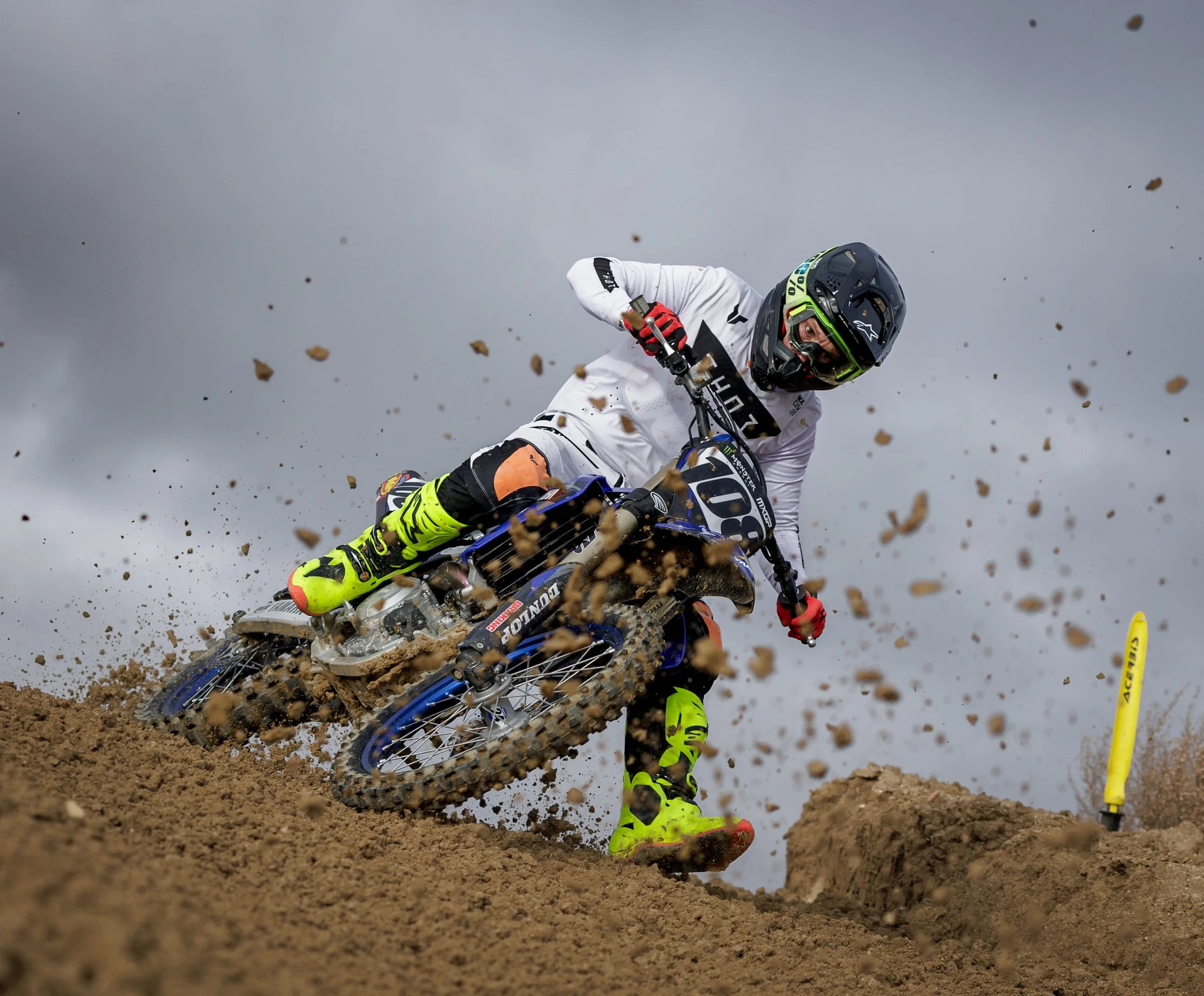 Un motociclista vestido con equipo de protección y ropa blanca, montando una moto de motocross azul, tomando una curva en un terreno de tierra, con un cielo nublado de fondo.