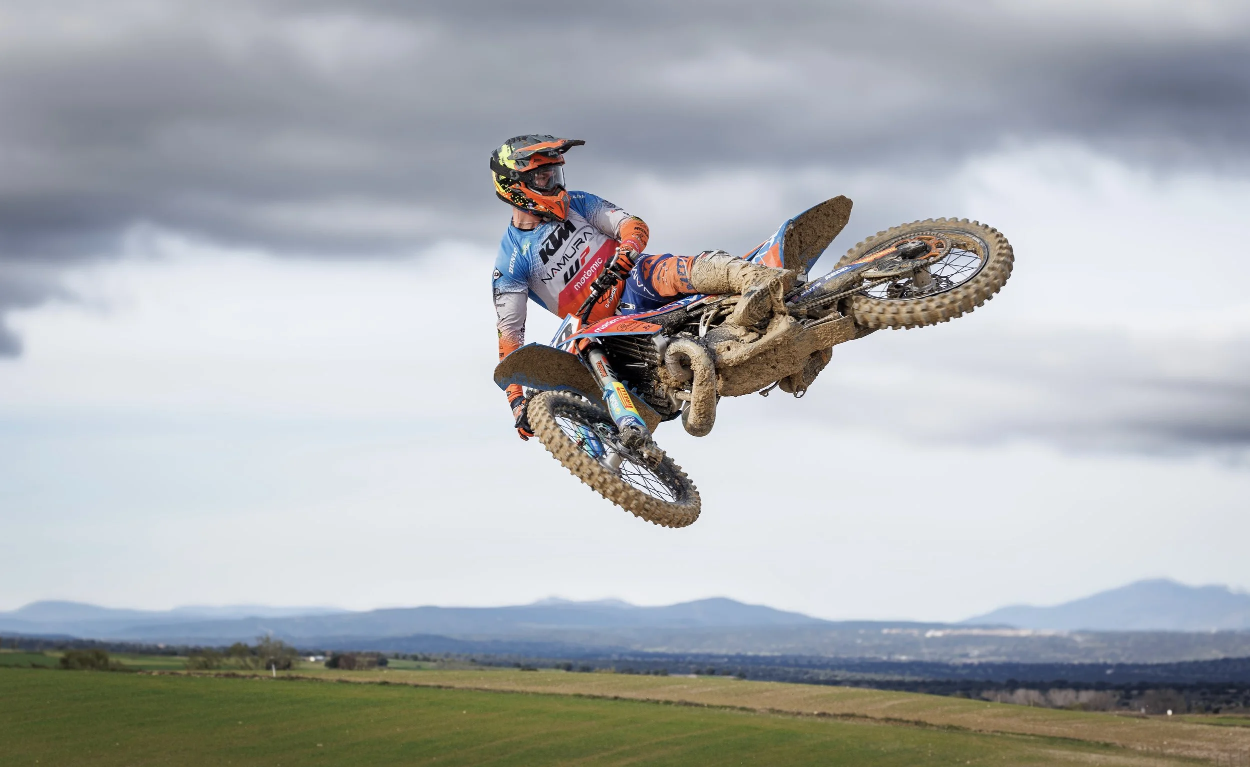 Piloto de motocicleta haciendo salto en una pista de campo, con cielo nublado y paisaje rural al fondo.