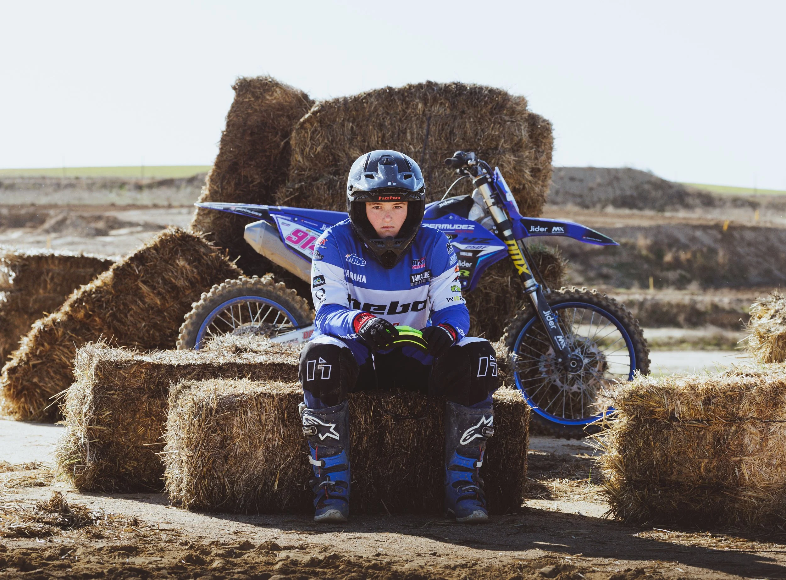 Joven piloto de motocross en ropa de competición, con casco, sentado sobre un bloque de heno, con una moto de dirt bike azul detrás en un campo al aire libre.