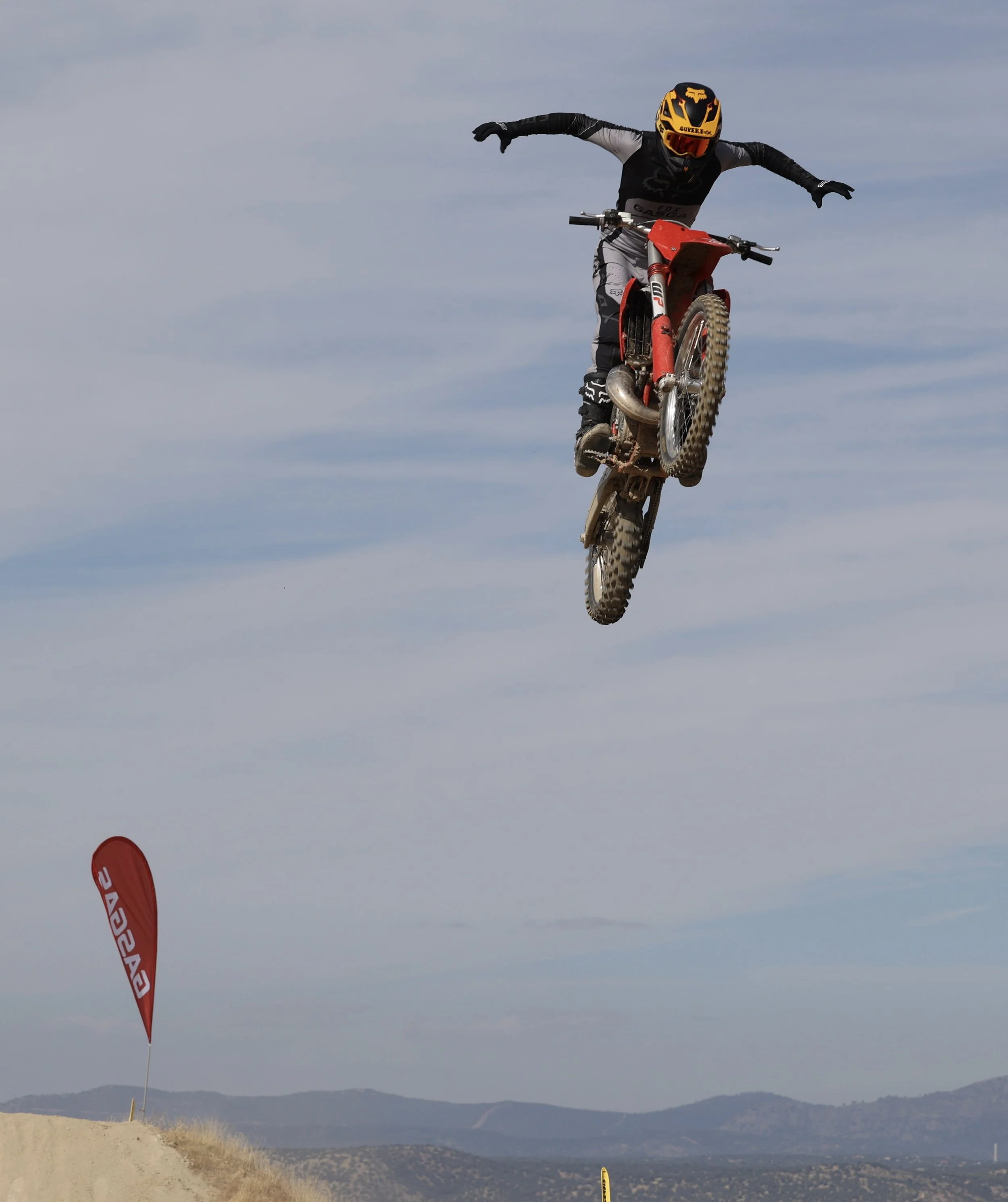 Un motociclista en vuelo sobre un terreno arenoso, usando casco y equipo de protección, durante una competencia de motocross.