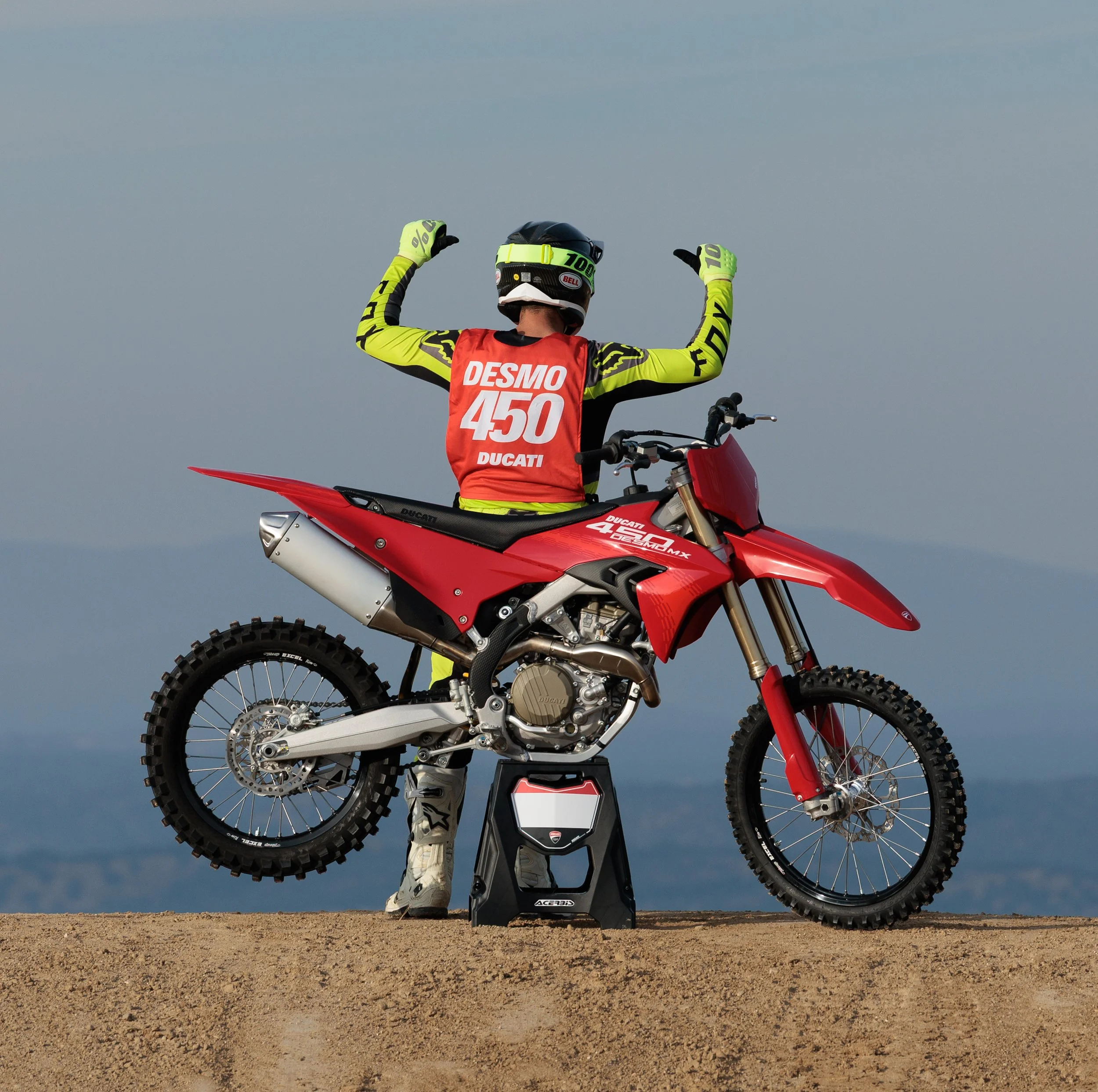 Un piloto de motocross en uniforme de competición, de espaldas, con brazos en alto, sentado en una motocicleta Ducati roja en un terreno de tierra, con un fondo de cielo y paisaje lejano.