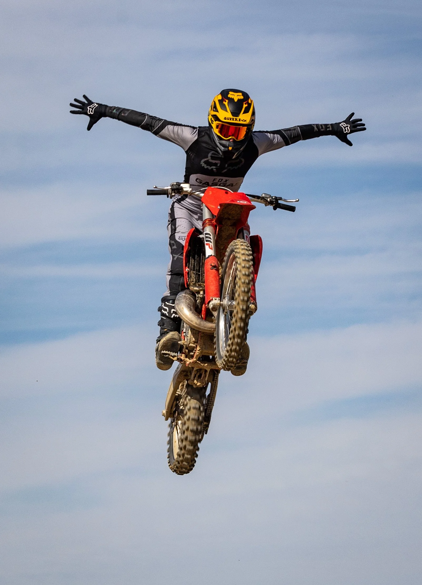 Persona en una motocicleta realiza un salto en el aire con brazos abiertos y fondo de cielo con nubes.
