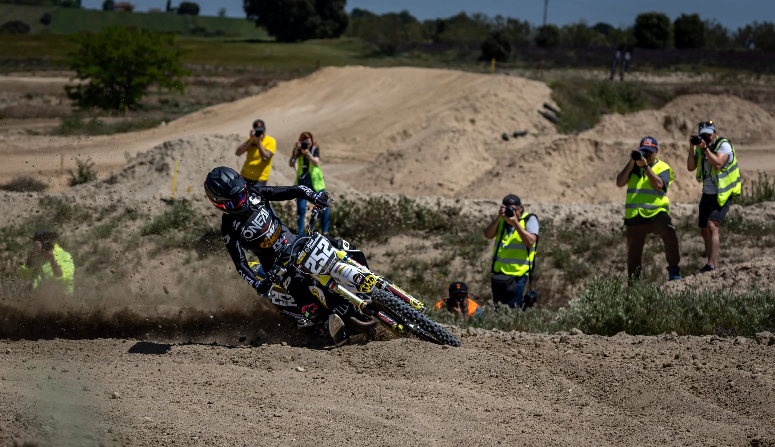 Un motociclista en un circuito de tierra durante una carrera de motocross, rodeado por varios fotógrafos y espectadores observando y tomando fotos.