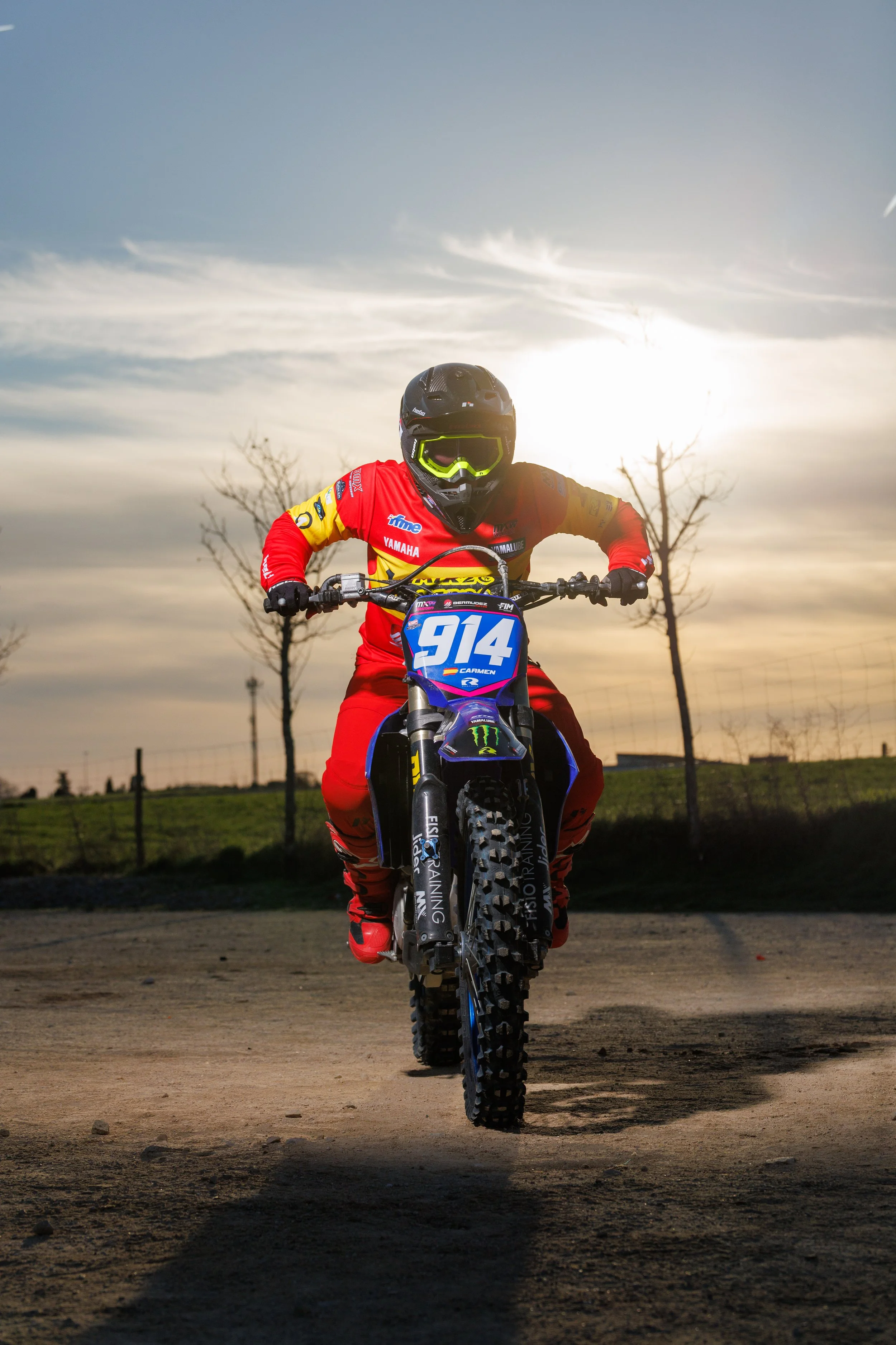 Un piloto de motocross en una moto, con casco y equipo de protección, en un terreno de tierra, con árboles y un cielo nublado de fondo.