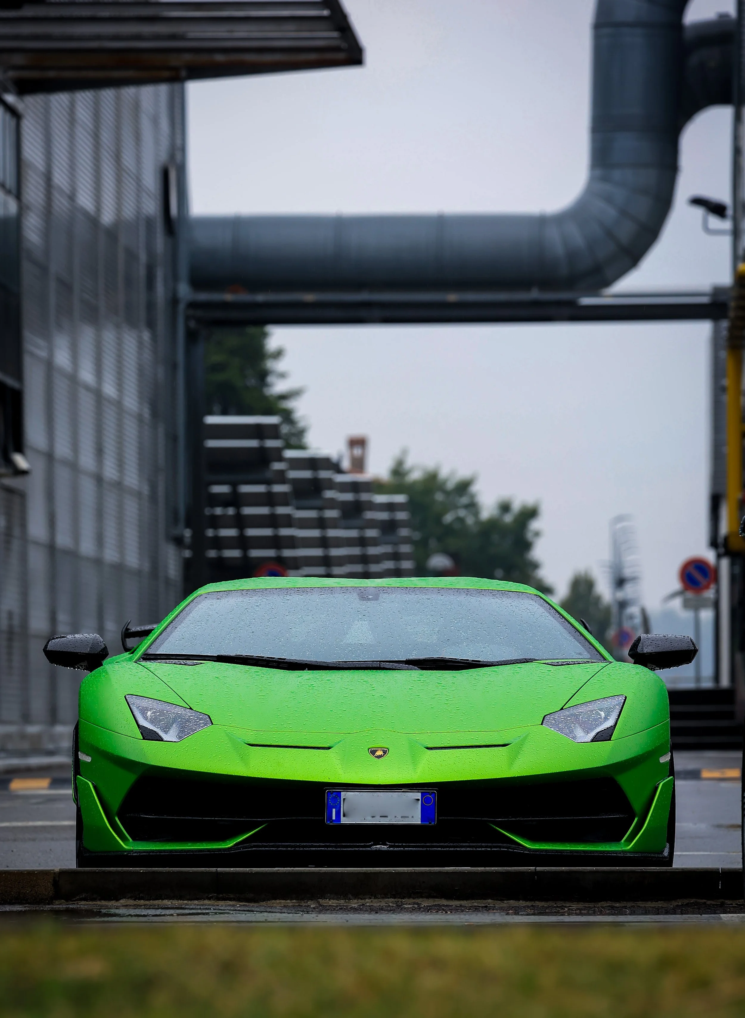 Coche deportivo verde estacionado en la calle, con edificios y tuberías en el fondo, día lluvioso.