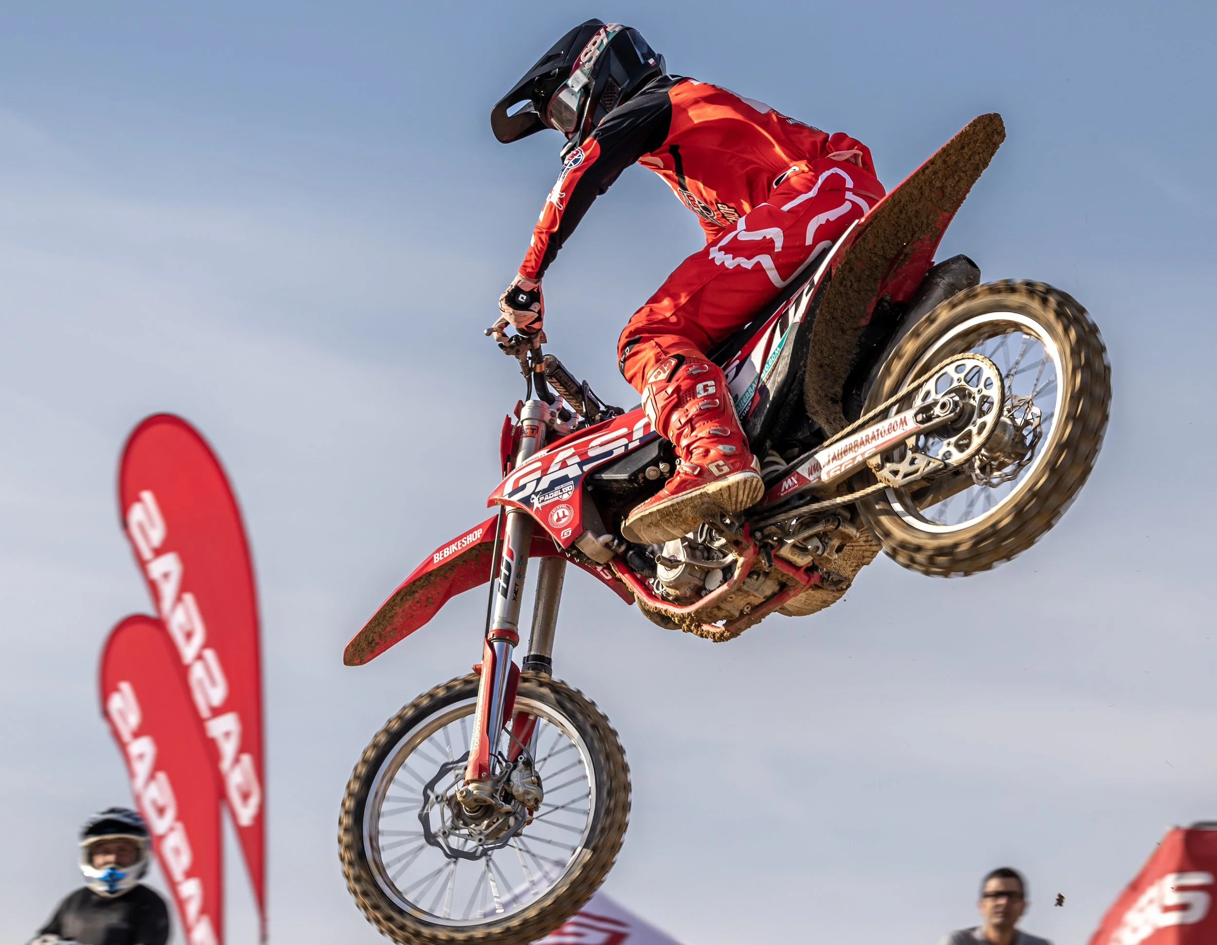 Un piloto de motocross en salto aéreo en un circuito al aire libre, con un traje rojo y un casco negro, en medio de una competencia.