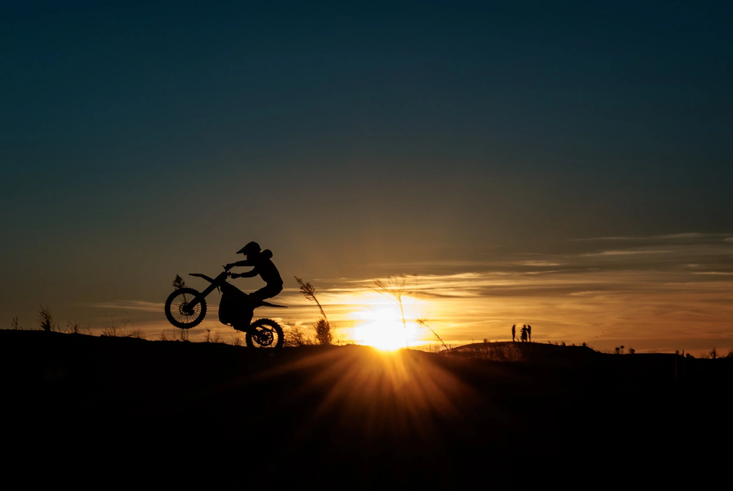 Persona haciendo acrobacias en bicicleta durante el atardecer en un campo abierto, con otras personas en el fondo.