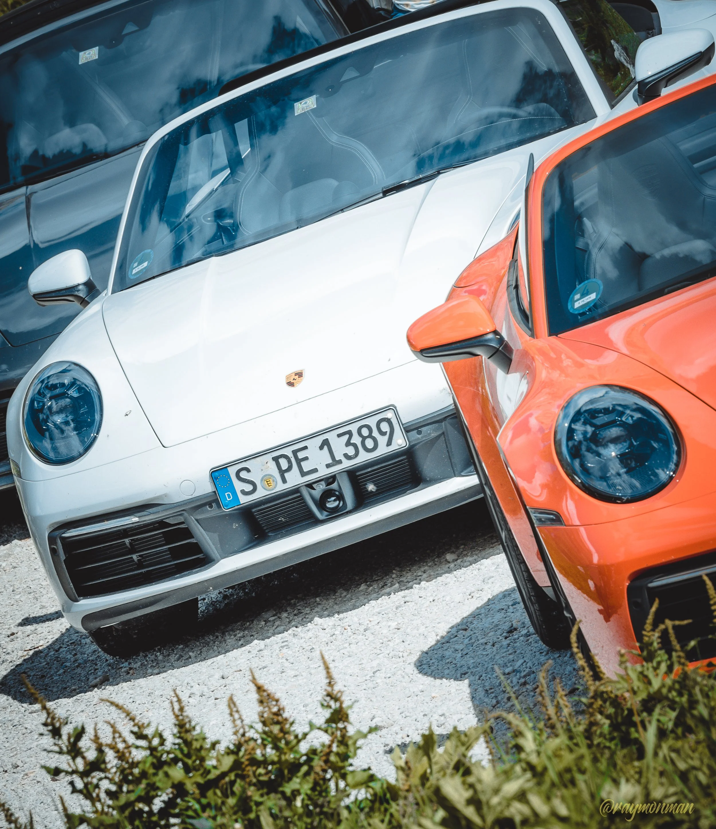Dos autos deportivos, un Porsche blanco y un Porsche naranja, estacionados en un aparcamiento con grava.