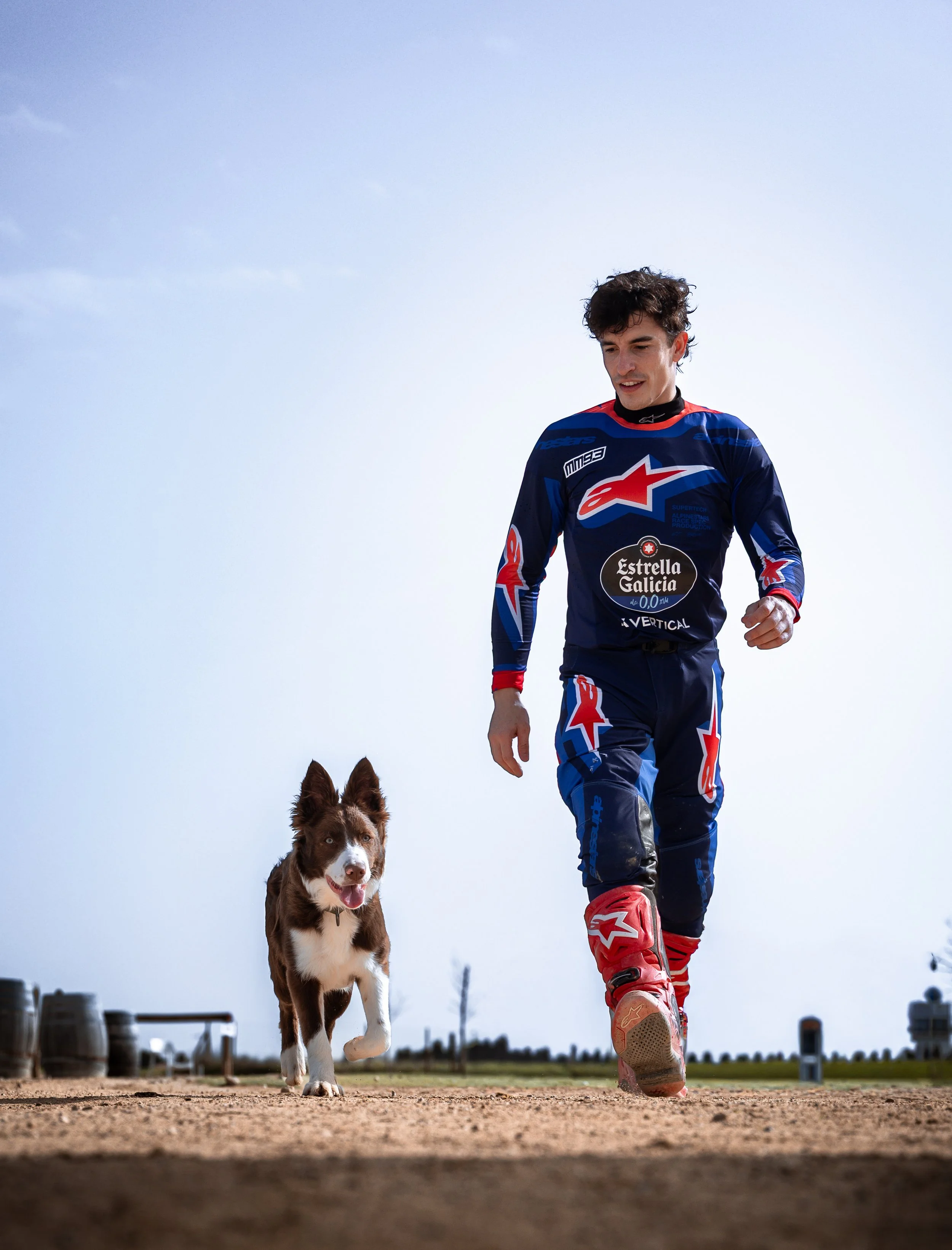 Joven hombre en ropa de motocross corriendo con un perro en un campo de tierra al aire libre bajo cielo despejado.
