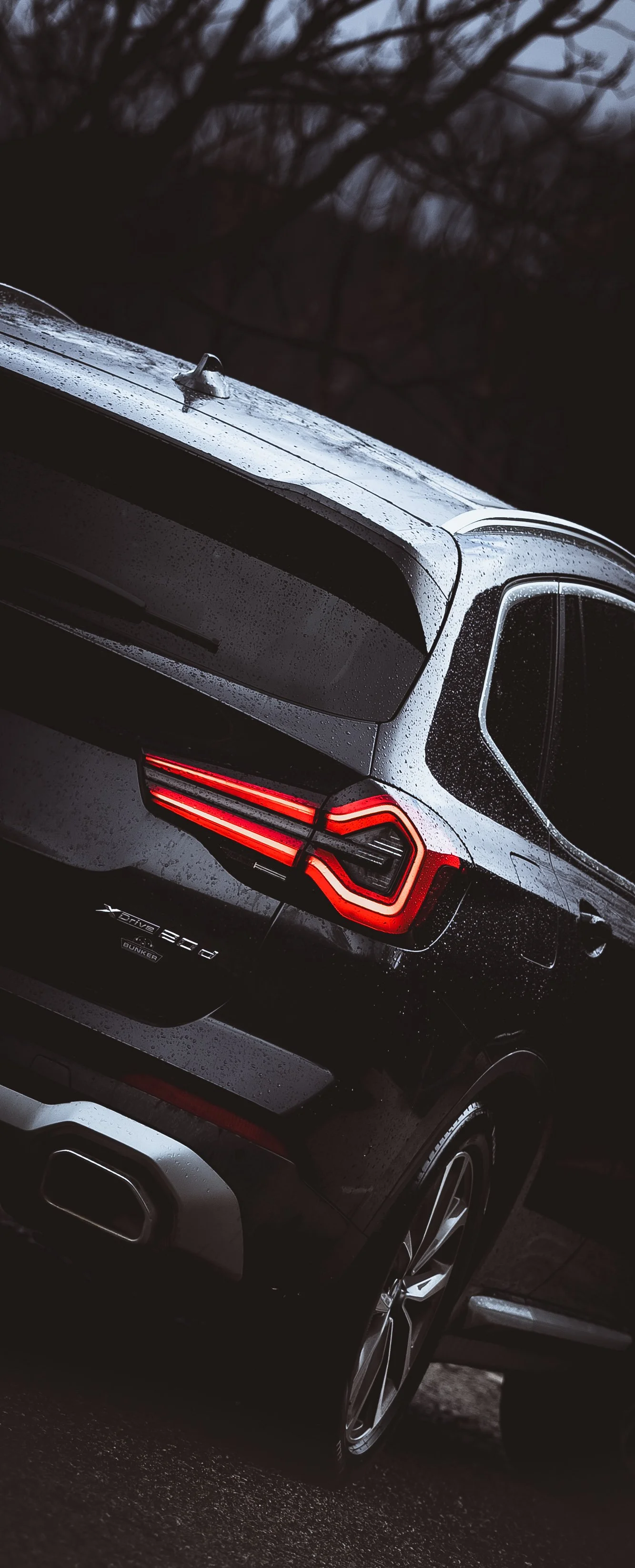 Parte trasera de un automóvil negro con luces LED rojas, en un ambiente oscuro con árboles y lluvia en la superficie del coche.