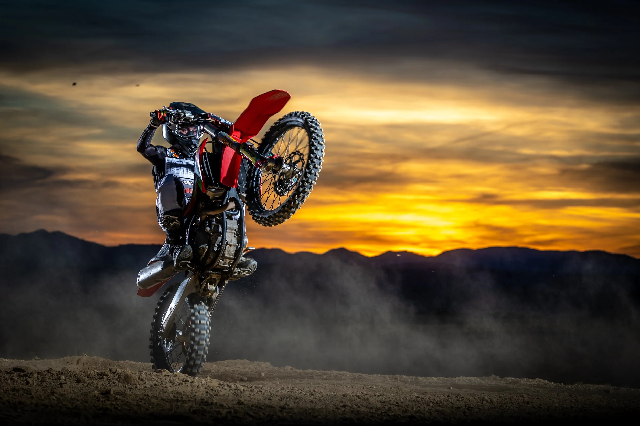 Un motero realizando un truco con una moto de motocross en un paisaje con montañas y un atardecer. Wheelie Sunset 