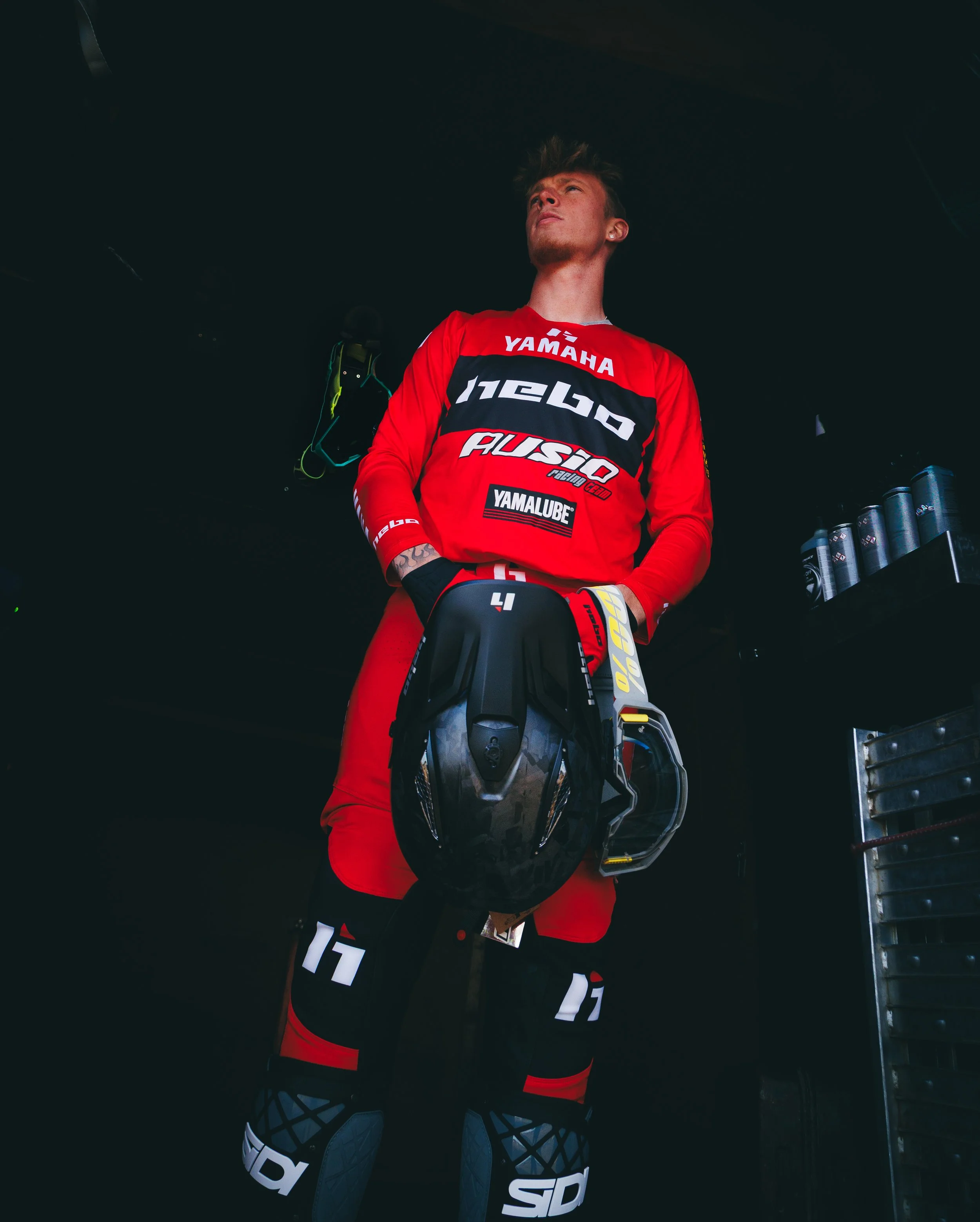 Joven piloto de motocross con traje y casco en la mano, de pie en un lugar oscuro, con estantería con latas a la derecha.