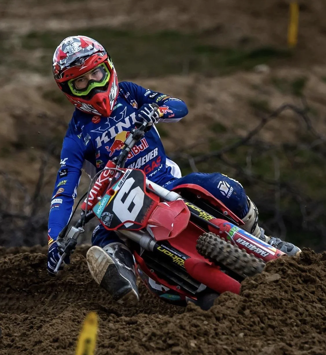Un piloto de motocross en una carrera, usando casco y equipo de protección, montado en una motocicleta roja en un terreno de tierra.
