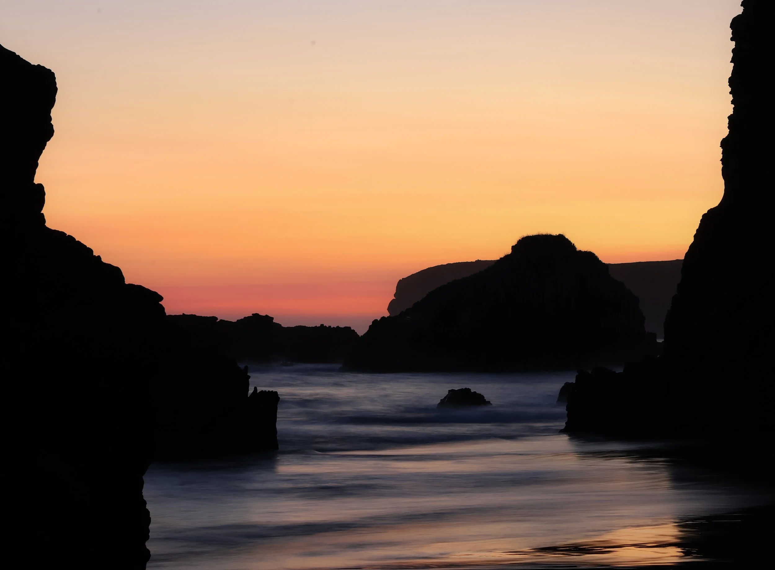 Atardecer en la playa con siluetas de rocas y reflejos en el agua.
