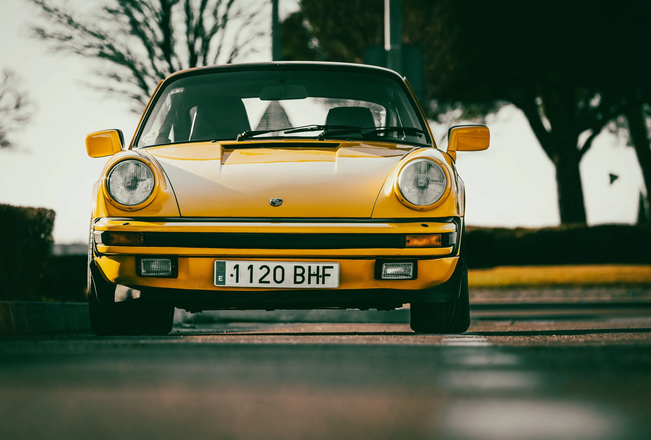 Coche deportivo clásico amarillo visto de frente en una calle con árboles en el fondo. Porsche