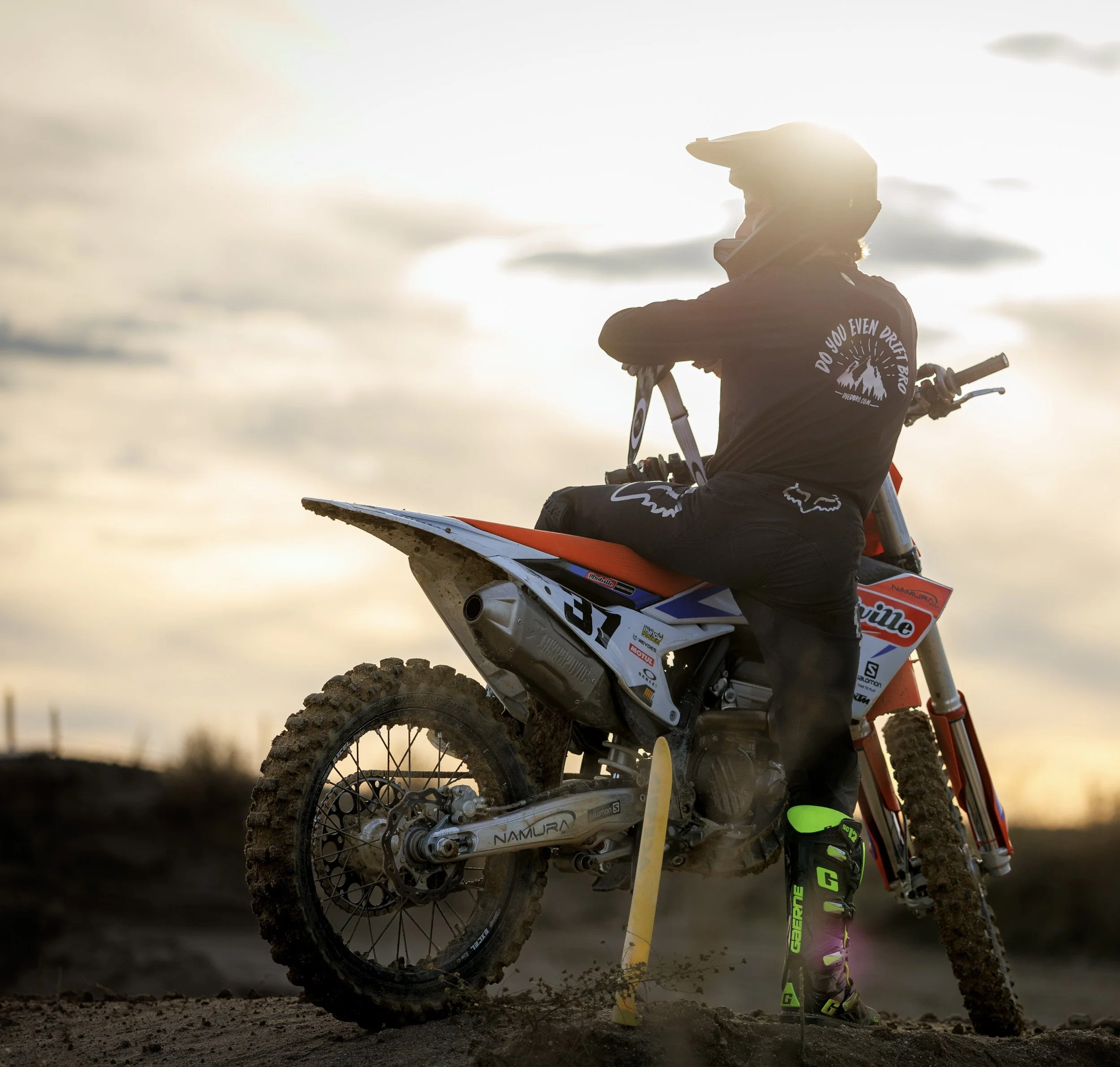 Piloto de motocross en la tierra durante atardecer, con moto y equipo de protección, sosteniendo un pie con el soporte de la moto.